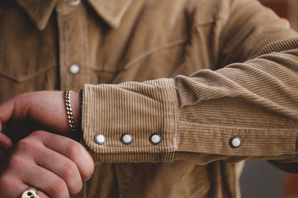 Close-up of a person's arm and hand adjusting the cuff of a tan corduroy button-up shirt. The shirt features three silver snap buttons on the cuff and a matching corduroy collar.