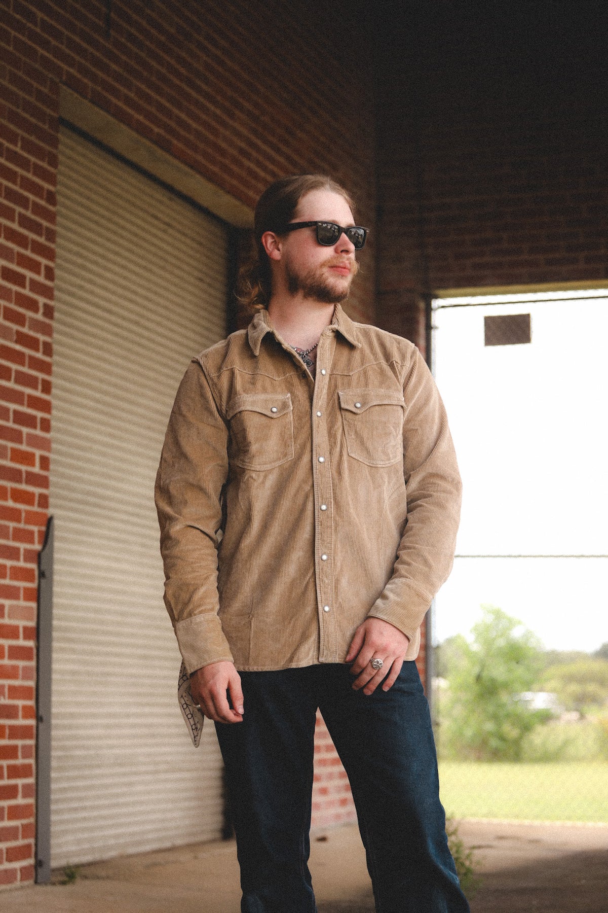 A man wearing a tan corduroy button-down shirt and dark jeans stands in a brick building under a metal roll-up door.