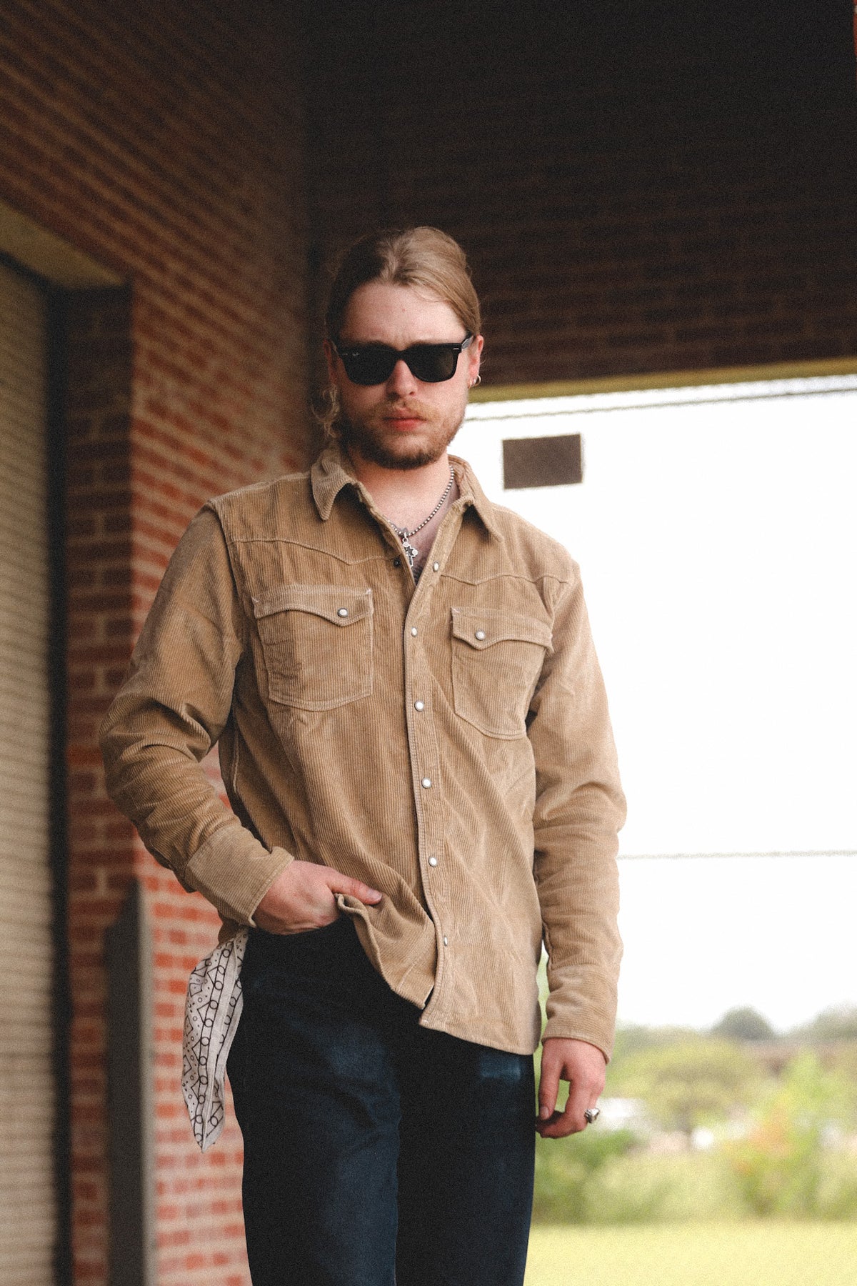 A man wearing a tan corduroy button-up shirt and dark jeans, standing in front of a brick building with a bright outdoor background.