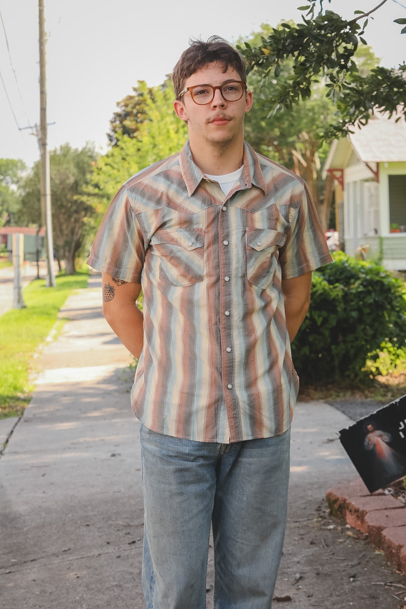 Man wearing a patterned shirt and glasses standing on a sidewalk with trees and houses in the background.