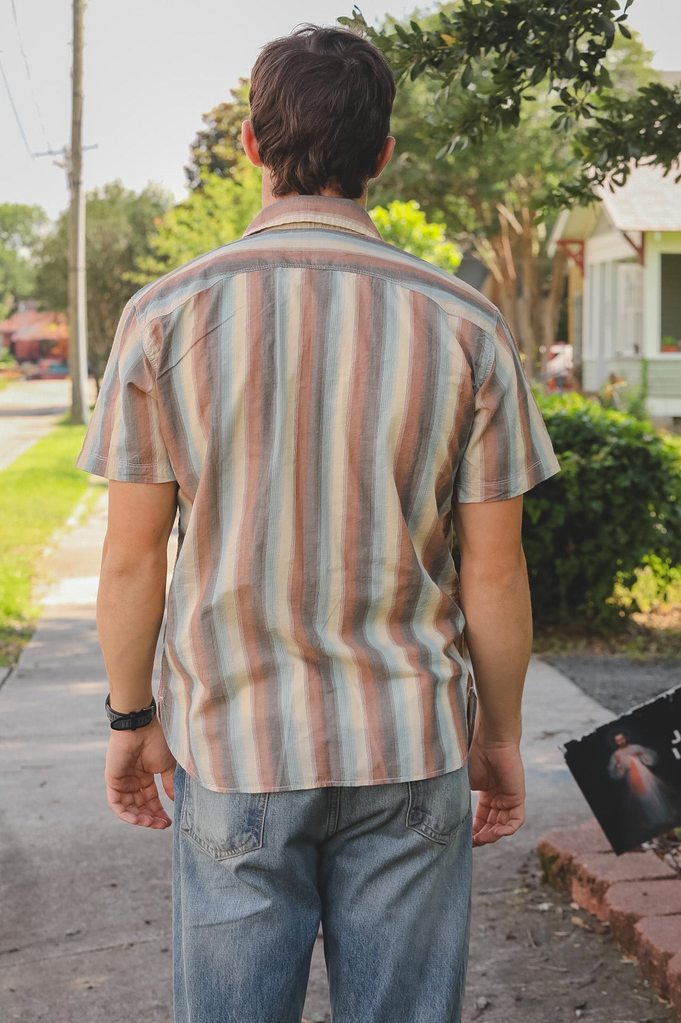 Person wearing a striped shirt and jeans walking on a sidewalk with trees and houses in the background.