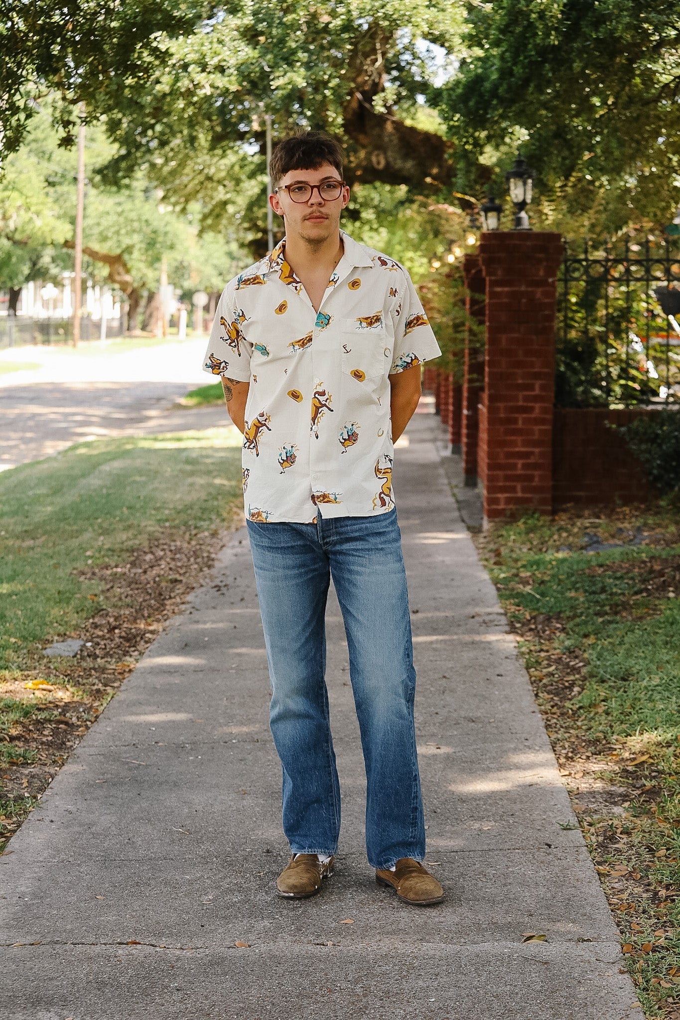 A man wearing a white short-sleeve button-up shirt with a colorful all-over print, blue jeans, and brown shoes, standing on a paved sidewalk outdoors.