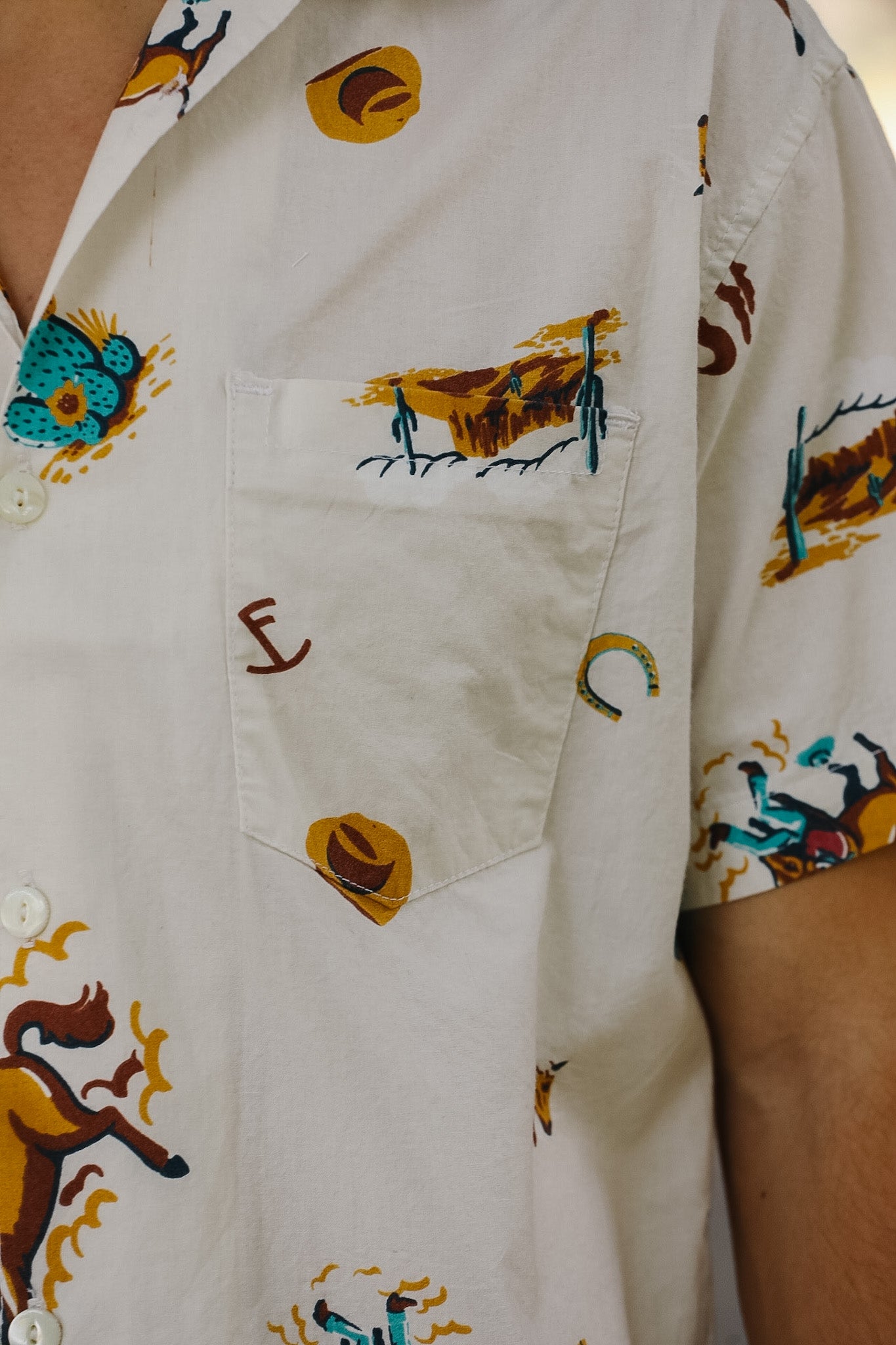 Close-up of a white short-sleeve button-up shirt with a chest pocket, featuring a colorful printed pattern of horses, cacti, and western motifs.