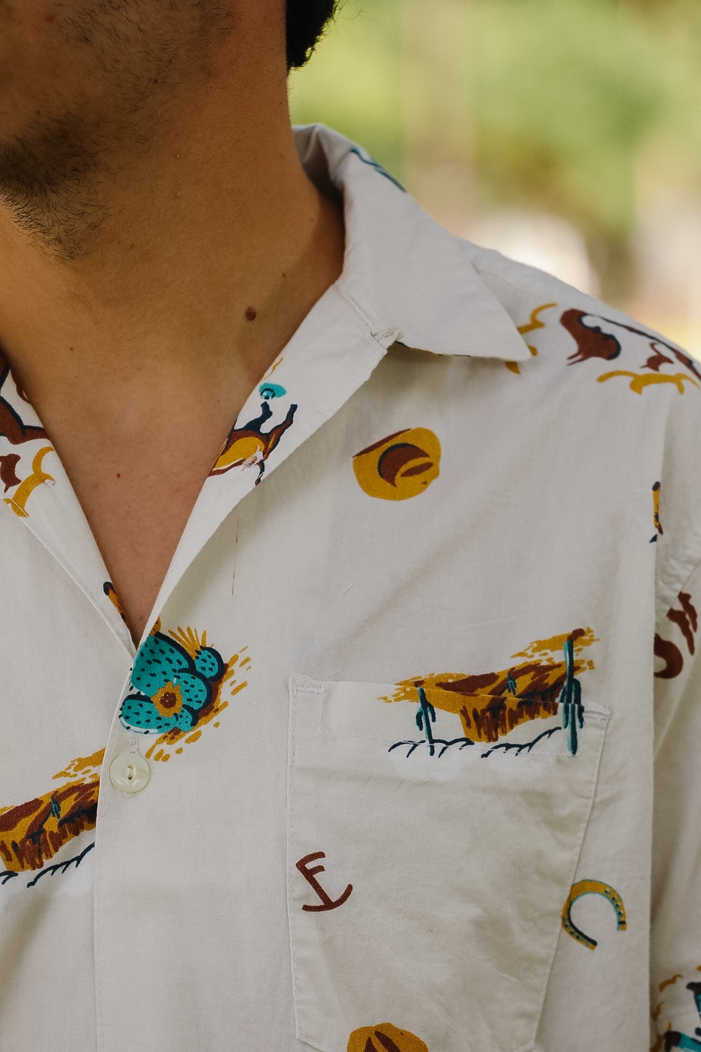 Close-up of a man wearing a white short-sleeve button-up shirt with a colorful embroidered pattern of horses, boots, and buckles.