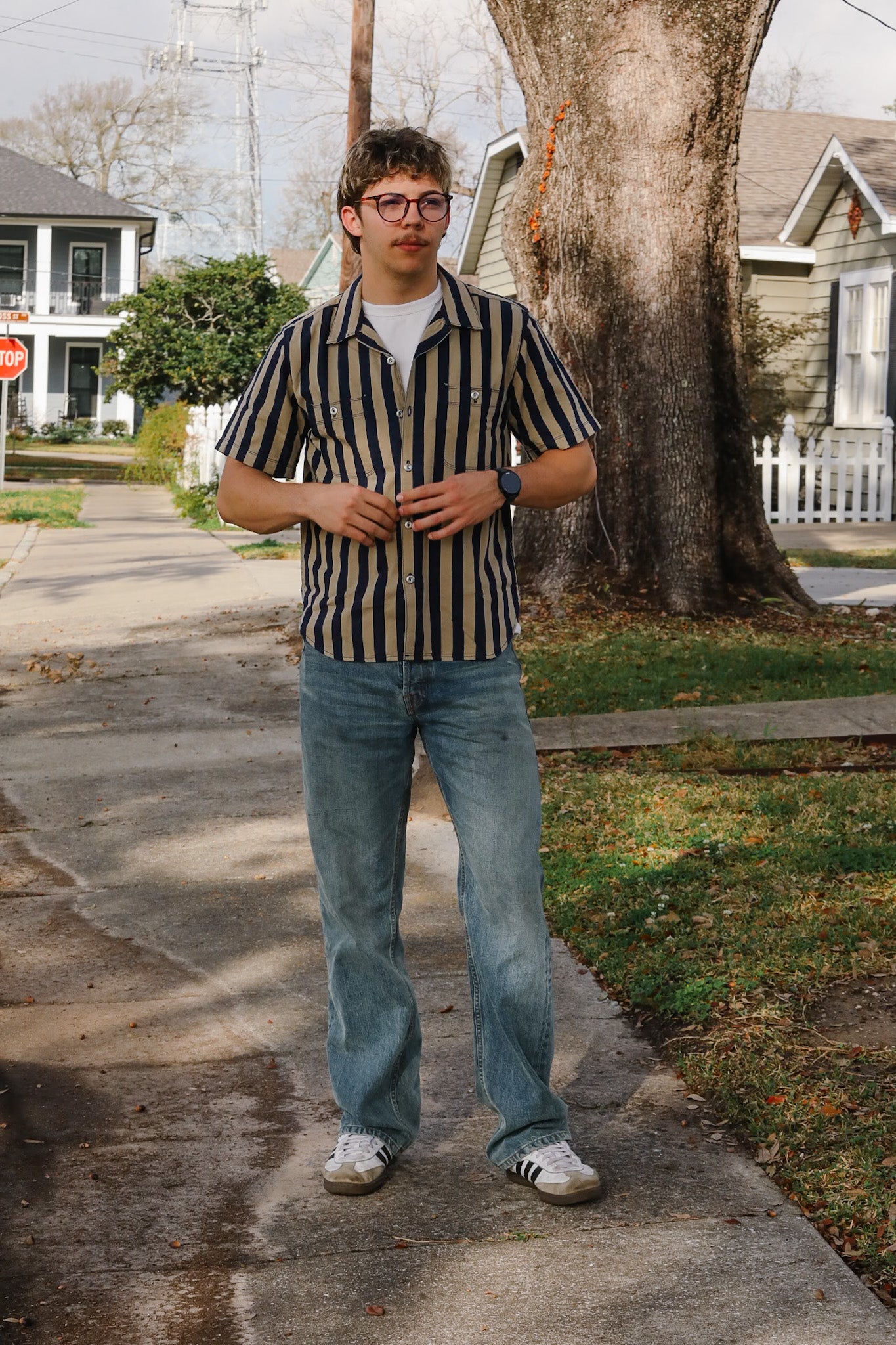 Man in a striped shirt and jeans standing on a sidewalk with houses and trees in the background