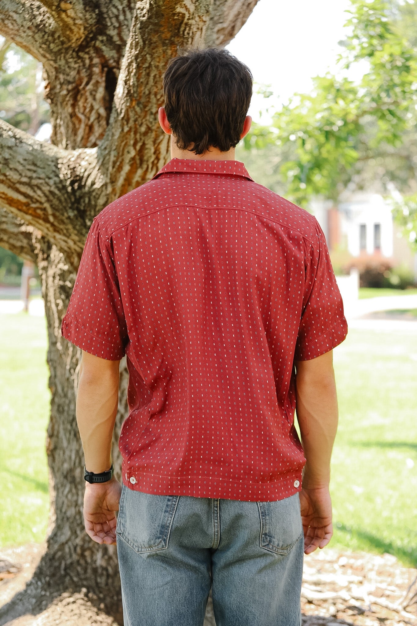 Person wearing a red patterned shirt standing next to a tree in an outdoor setting