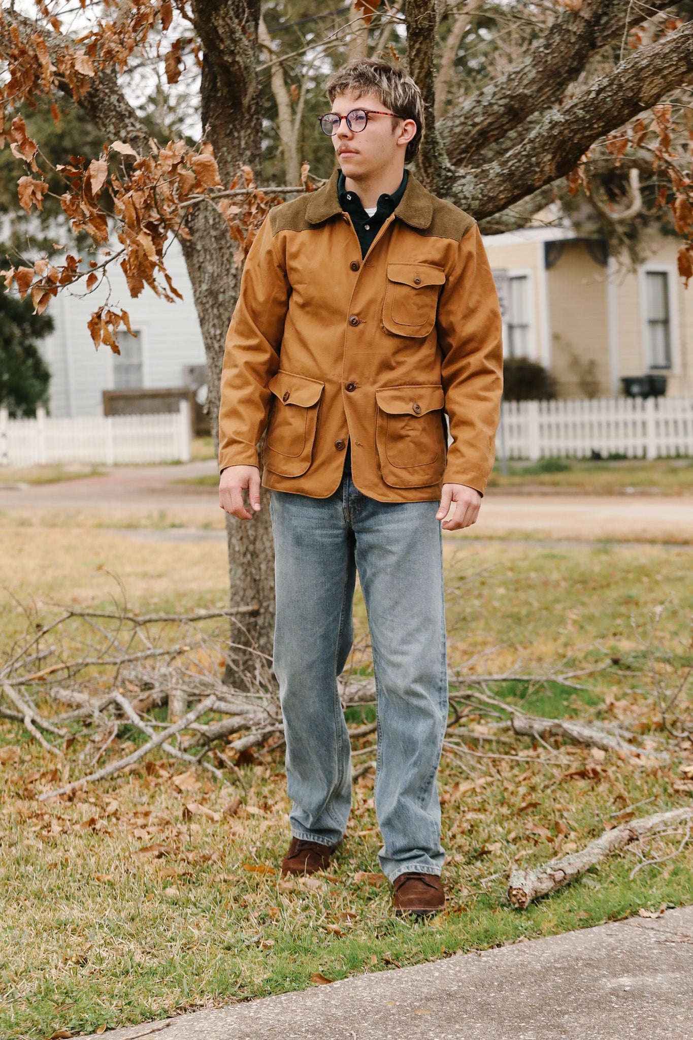 Man wearing a brown jacket and glasses standing outdoors near a tree