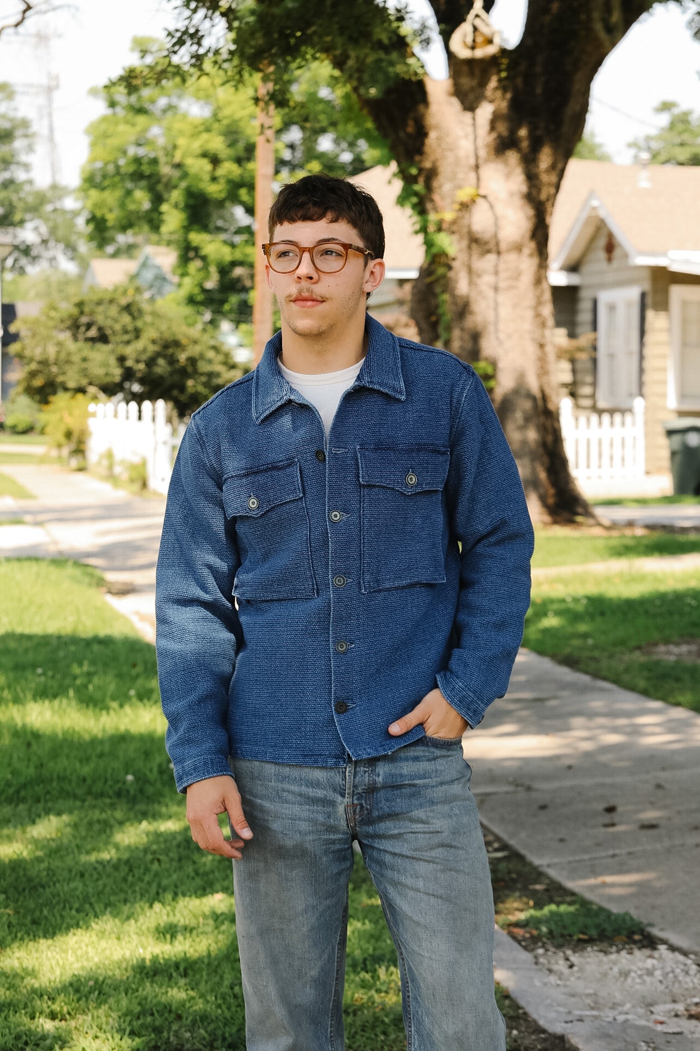 Man wearing a blue denim jacket and jeans standing on a sidewalk with trees and houses in the background.