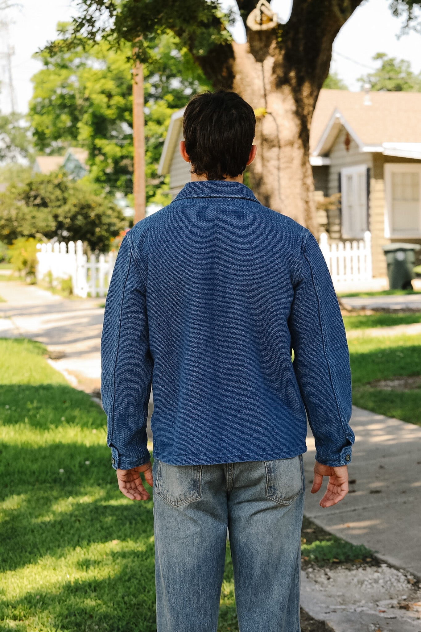 Person wearing a blue jacket and jeans walking on a sidewalk with a house and trees in the background