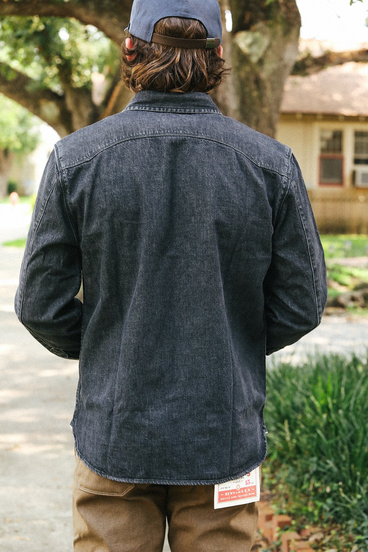 Person wearing a dark denim jacket and cap, standing outdoors with trees and a building in the background.
