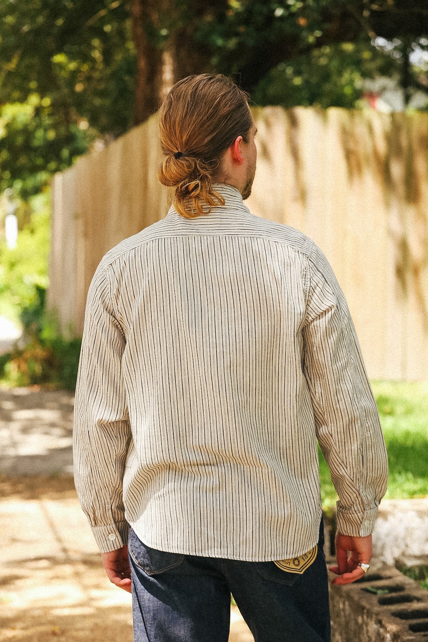 A man viewed from behind wearing a beige and brown vertically striped long-sleeve button-down shirt and dark blue jeans, standing outdoors in front of a wooden fence and trees.