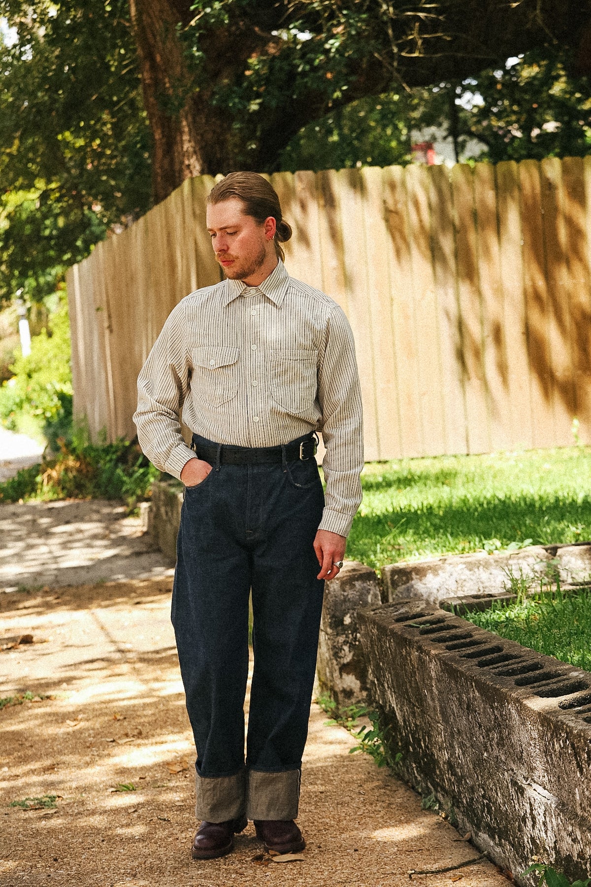 A man standing outdoors on a dirt path wearing a light beige vertically striped long-sleeve button-down shirt, dark blue cuffed jeans, and brown boots, with a wooden fence and trees in the background.