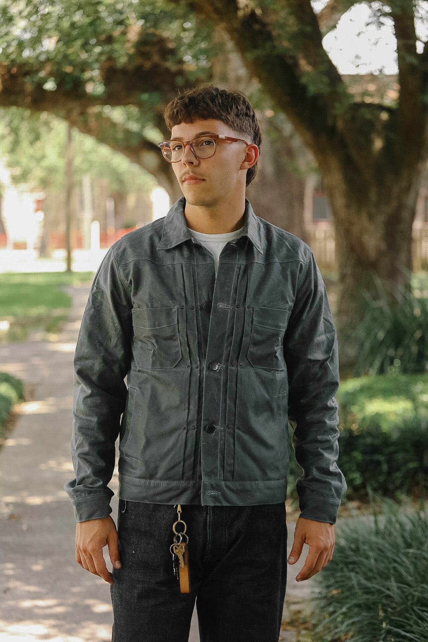 A man wearing a dark gray utility jacket, dark jeans, and glasses, standing on a tree-lined path.
