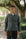 A man wearing a dark gray utility jacket, dark jeans, and glasses, standing on a tree-lined path.