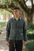 A man wearing a dark gray utility jacket, dark jeans, and glasses, standing on a tree-lined path.