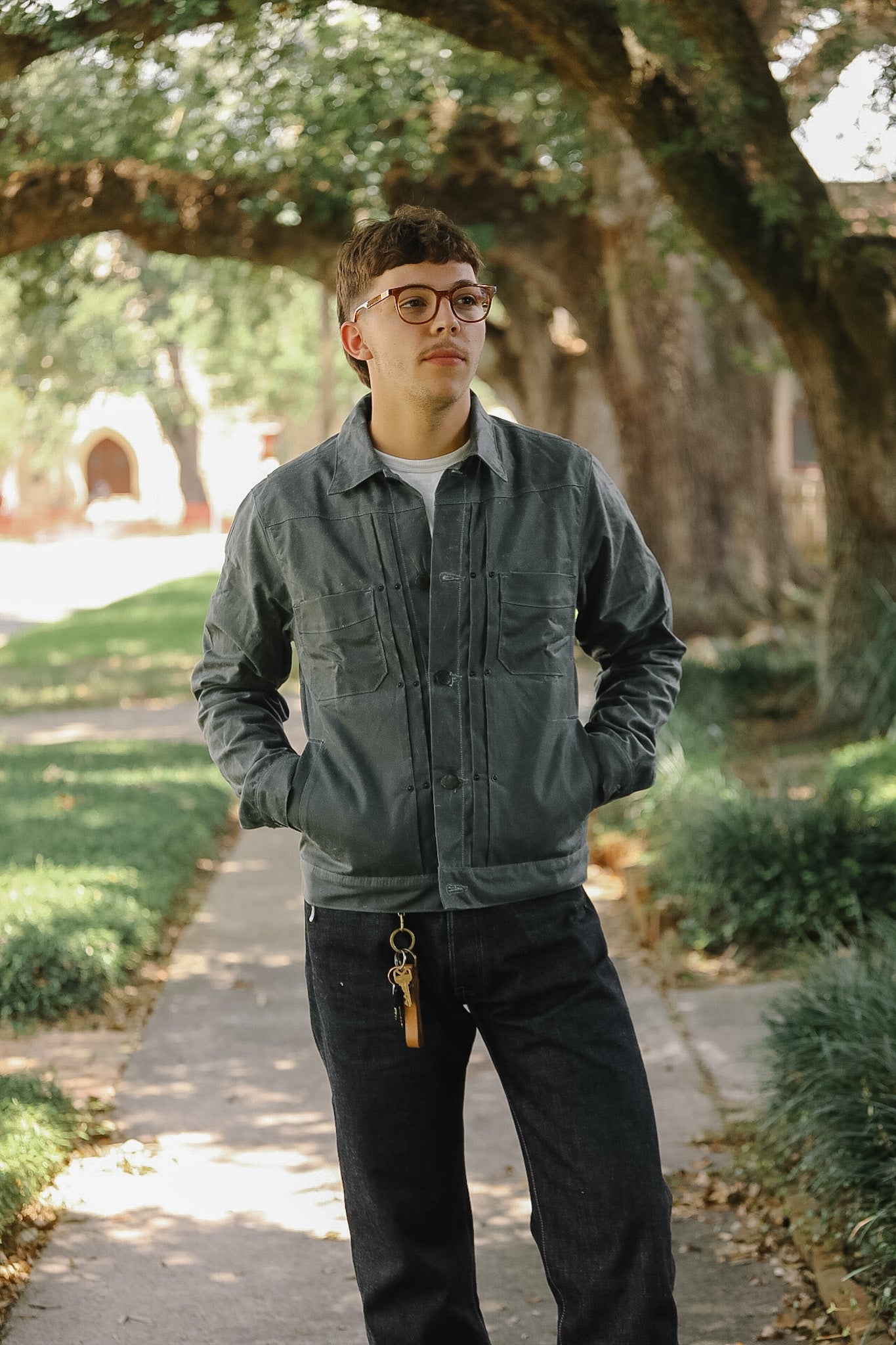 A young man wearing a dark gray utility jacket, black jeans, and glasses stands on a tree-lined path with his hands in his pockets.