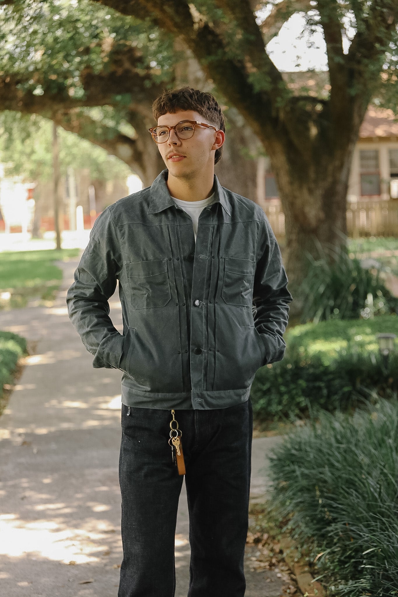 A man wearing a dark gray utility jacket, black jeans, and glasses stands on a tree-lined path with his hands in his pockets.