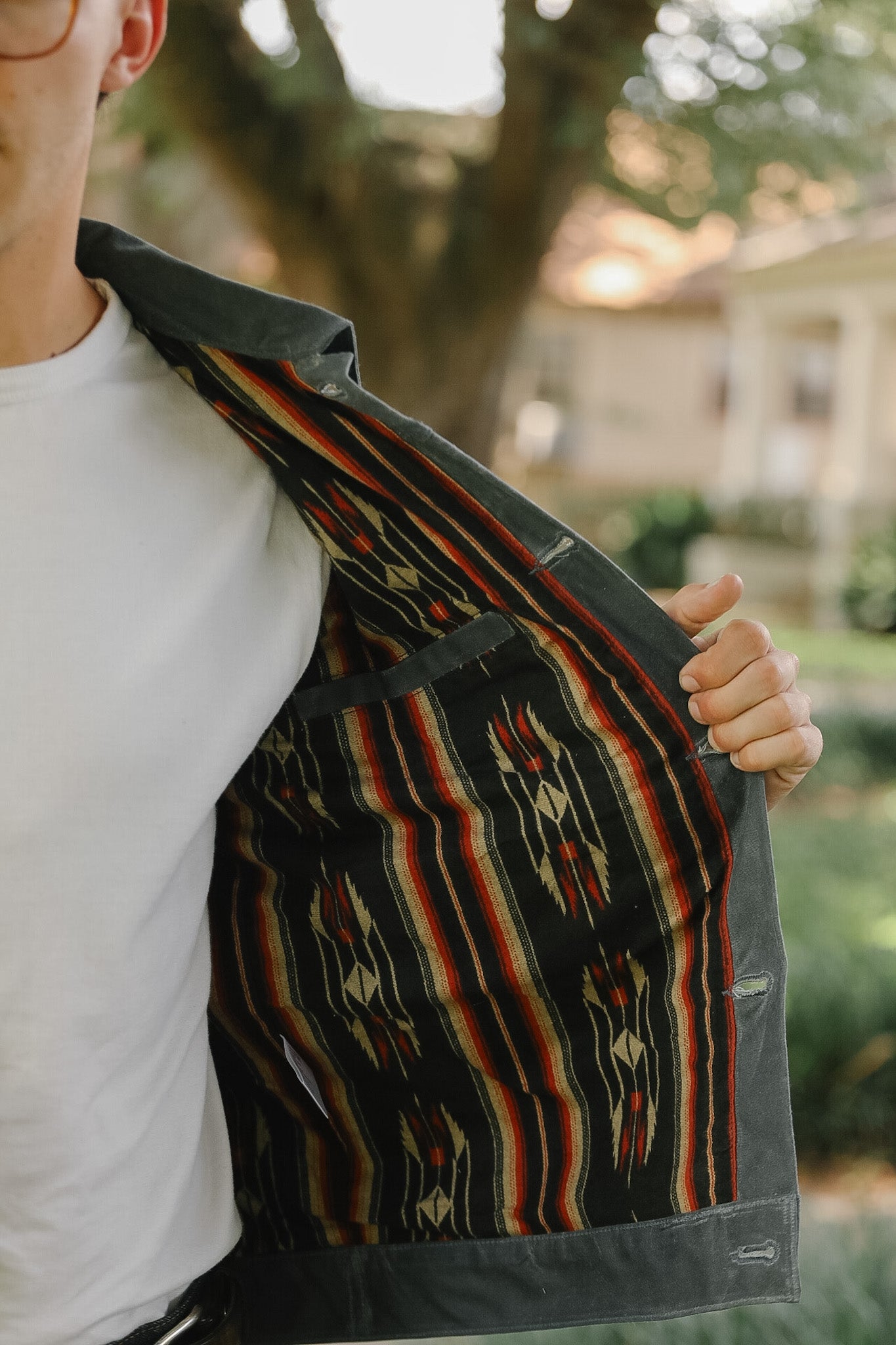 A person outdoors wearing a white t-shirt and holding open a dark gray jacket to show a colorful Southwestern-style striped patterned lining.