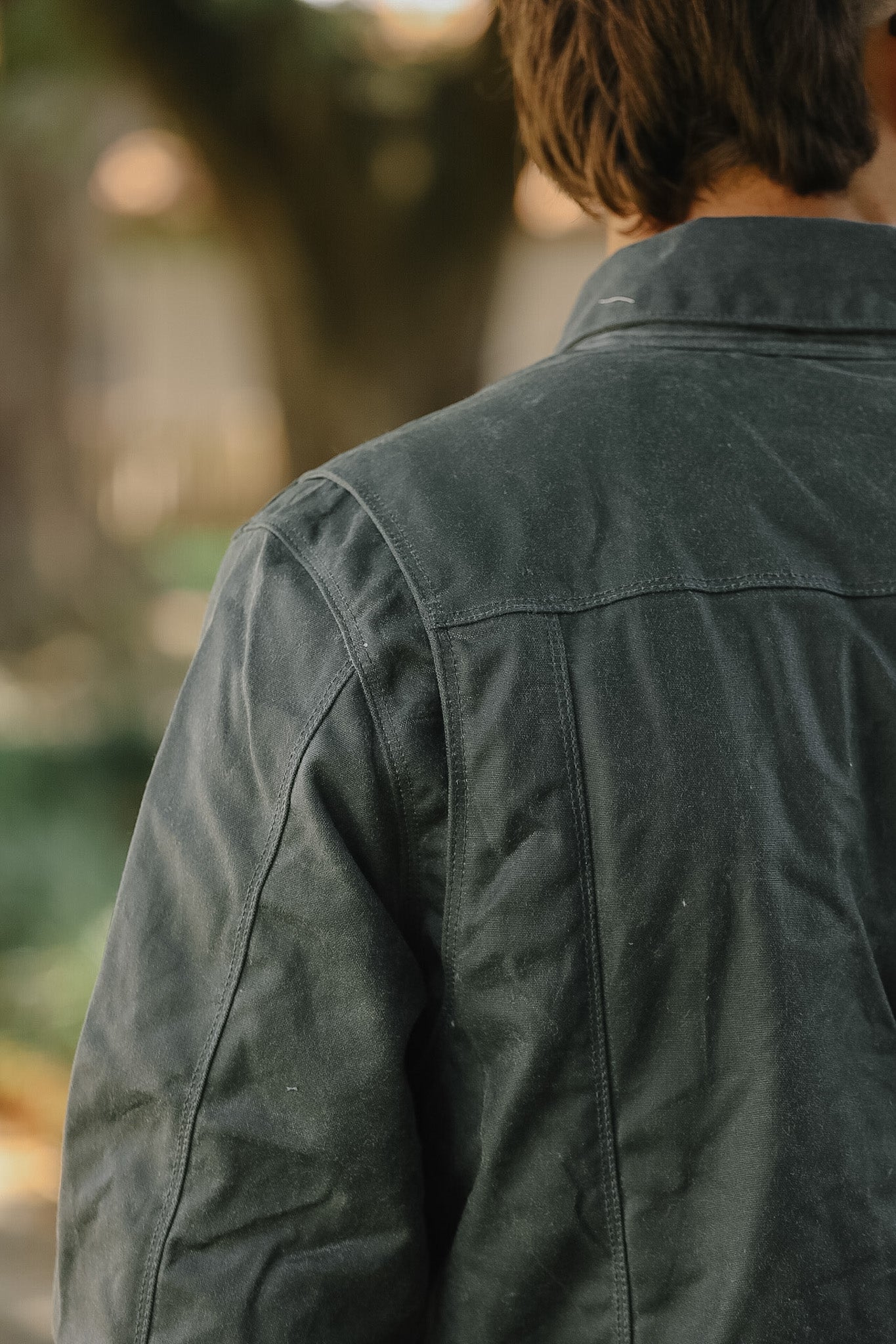 A close-up back view of a person wearing a dark charcoal gray utility jacket in an outdoor setting with a blurred background.