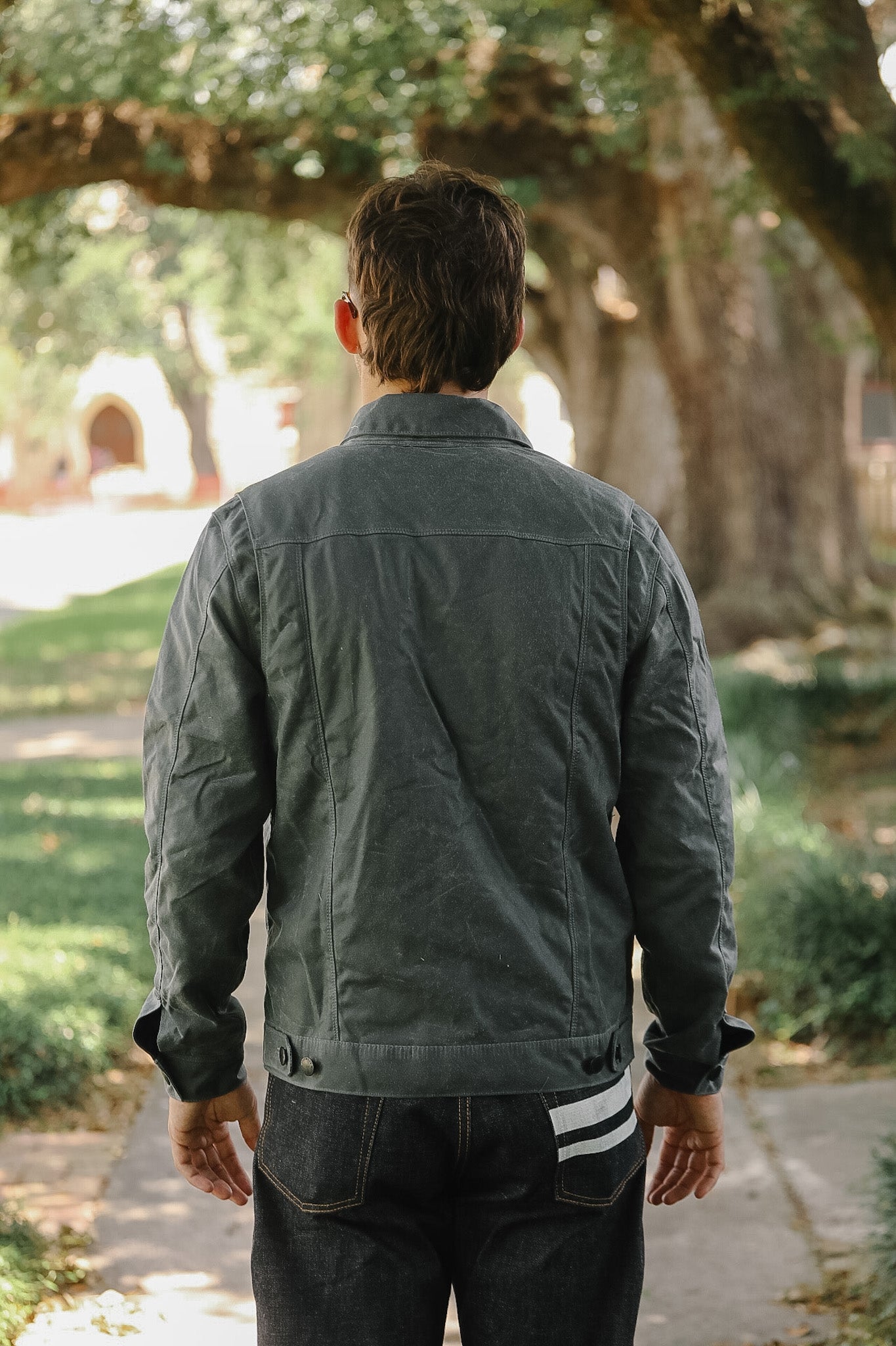 A man viewed from behind wearing a dark gray quilted jacket and dark jeans, standing on a paved path in a park with trees in the background.