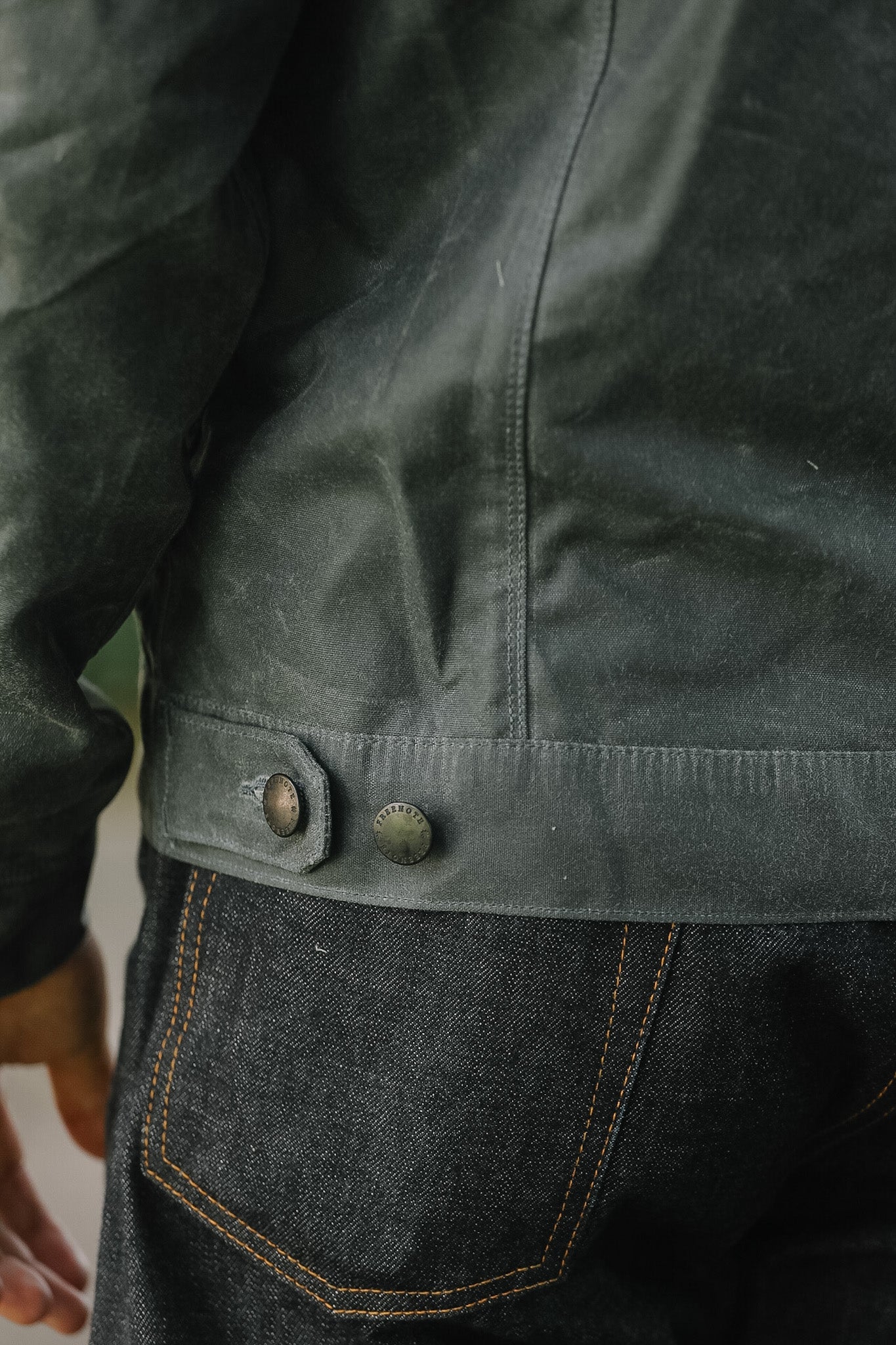 Close-up of a dark charcoal gray waxed canvas jacket worn over dark indigo jeans, showing the buttoned hem and fabric texture.