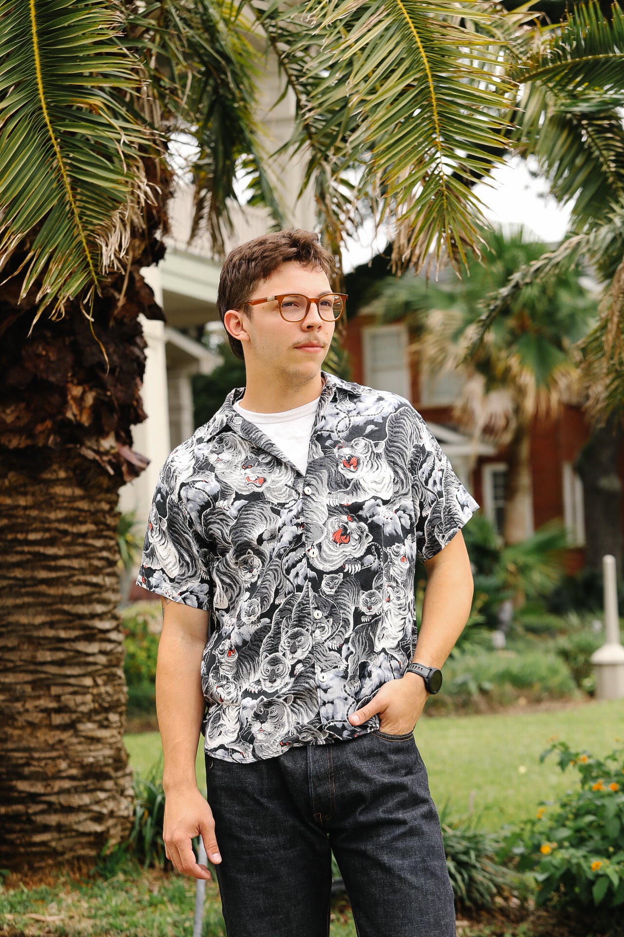 Man wearing a patterned shirt standing outdoors with palm trees and a house in the background