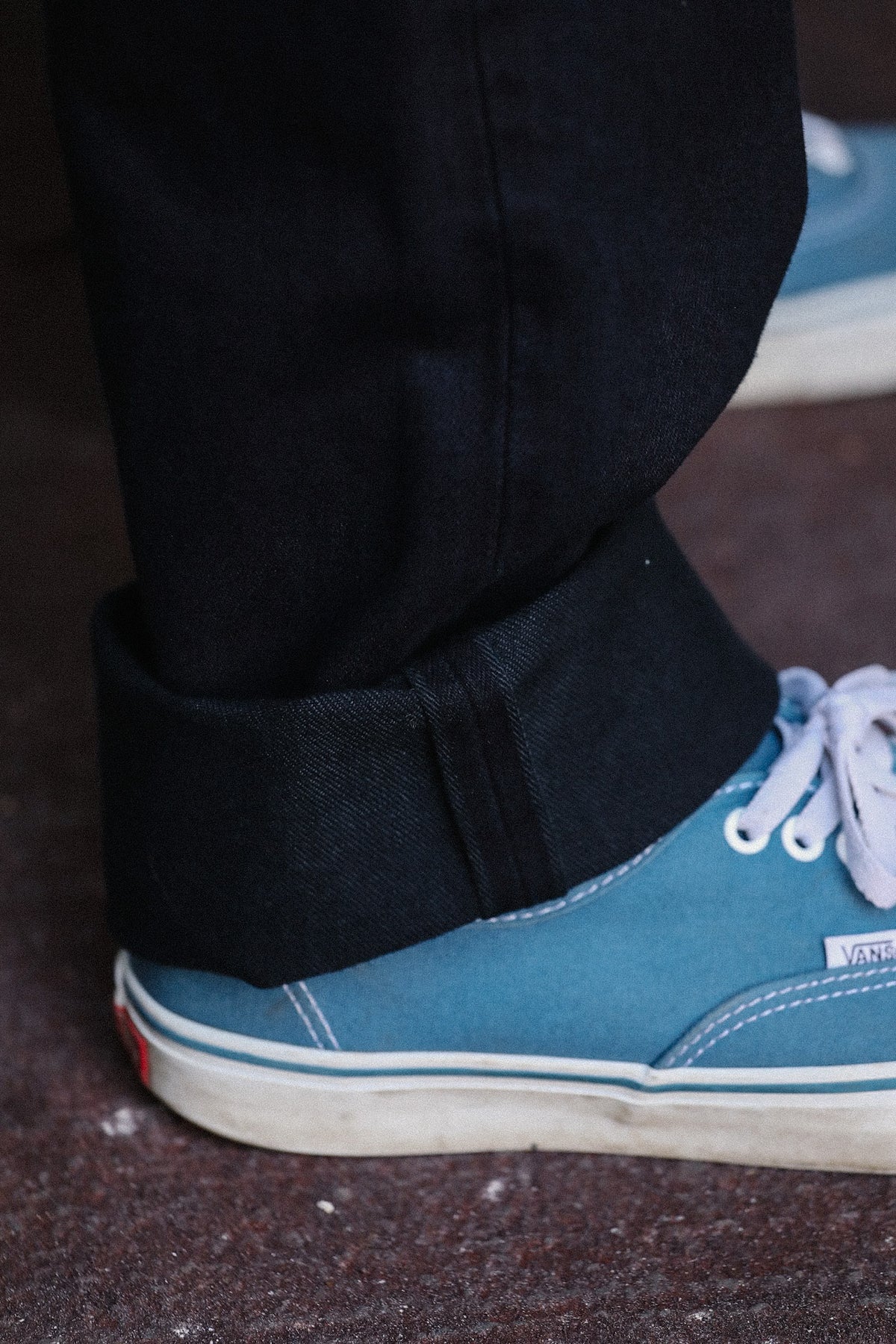 Close-up of a person's ankle wearing rolled-up black jeans and a light blue canvas sneaker with white laces, standing on a dark textured floor.