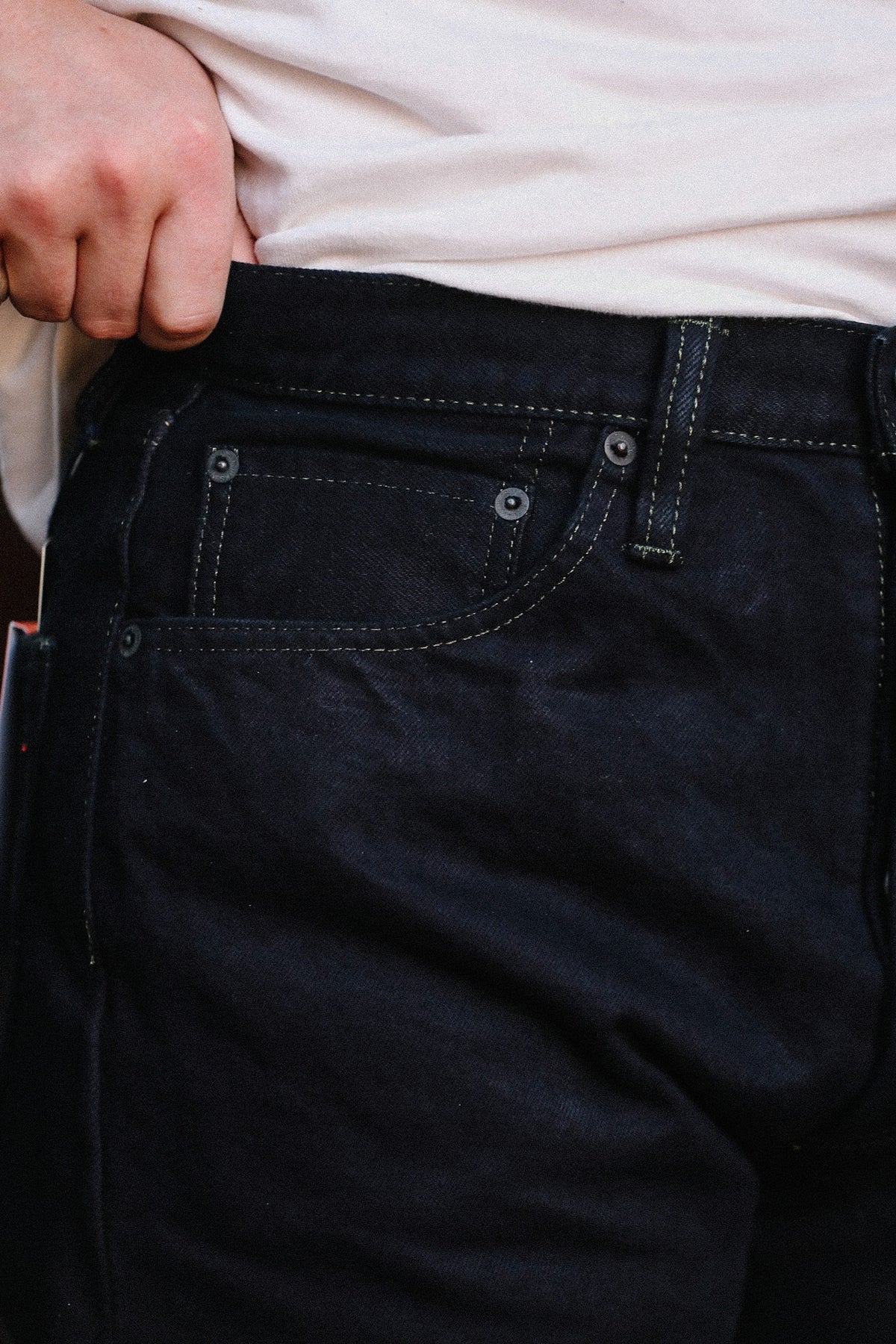 Close-up of a person wearing dark indigo denim jeans with a light-colored t-shirt, showing the waistband, belt loops, and front pocket details.