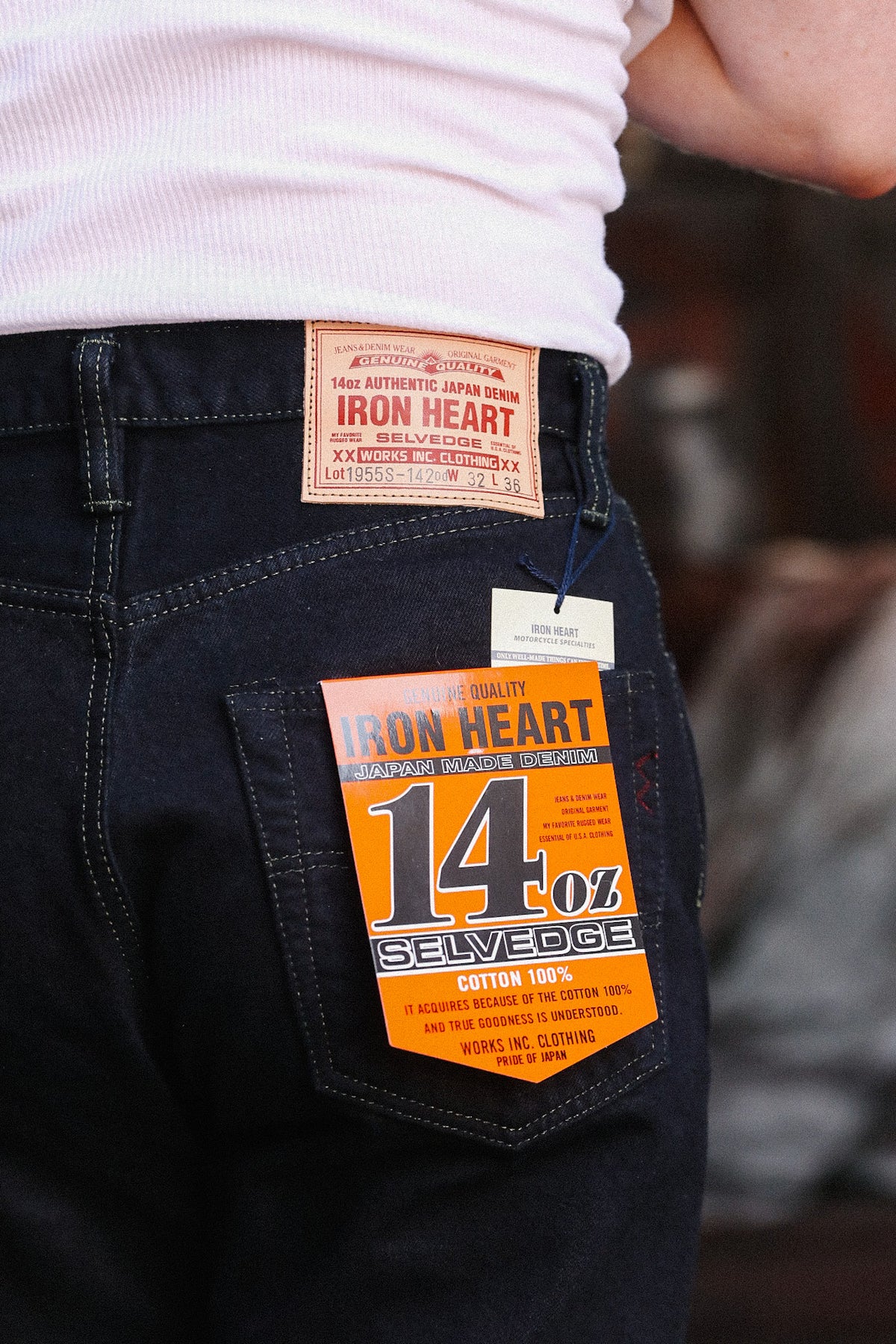 Close-up of the back pocket of dark indigo jeans with an orange Iron Heart 14oz Selvage paper tag attached, worn by a person in a white ribbed top.