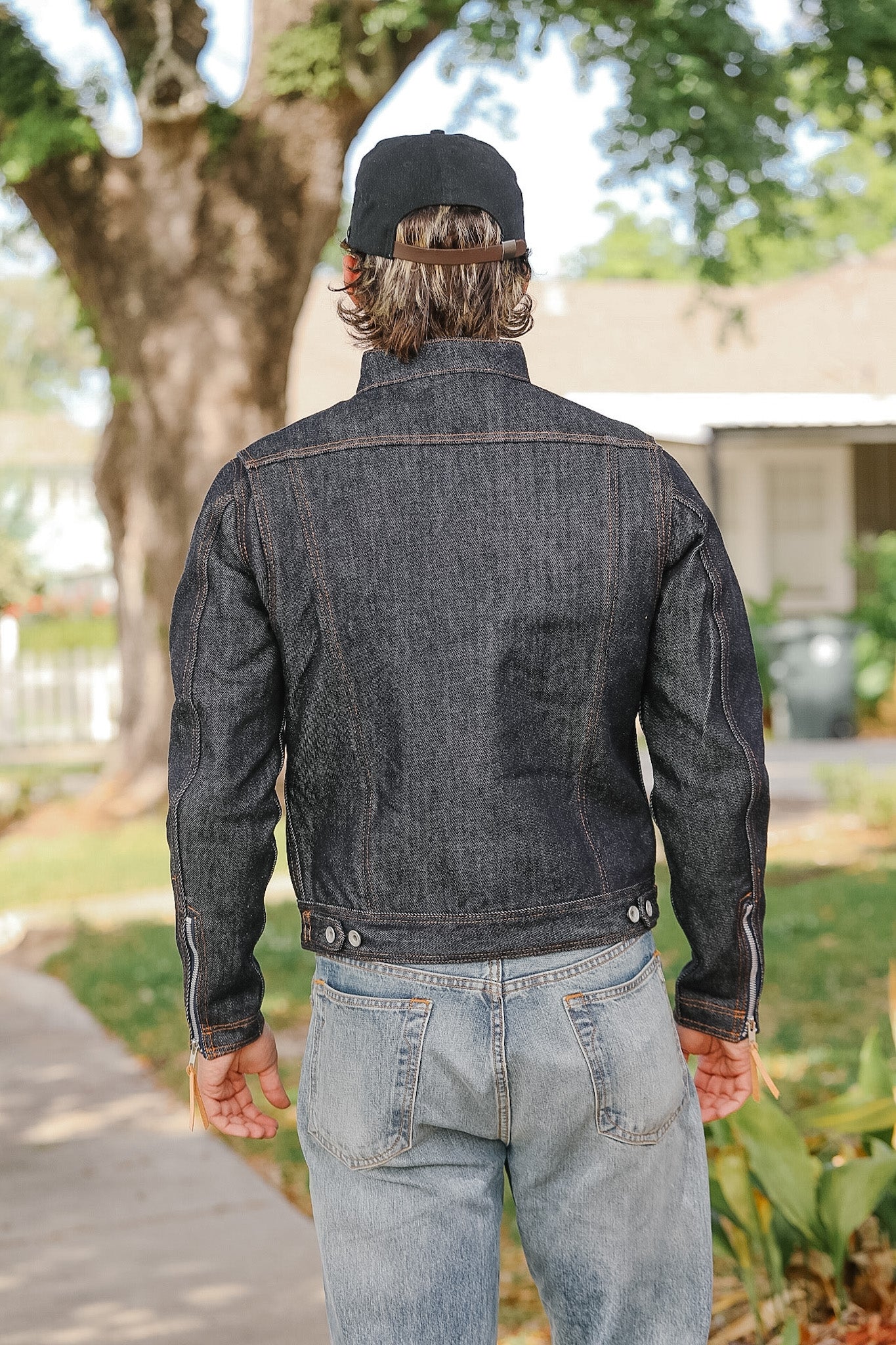 Person wearing a dark denim jacket and light blue jeans standing outdoors with trees and a house in the background.