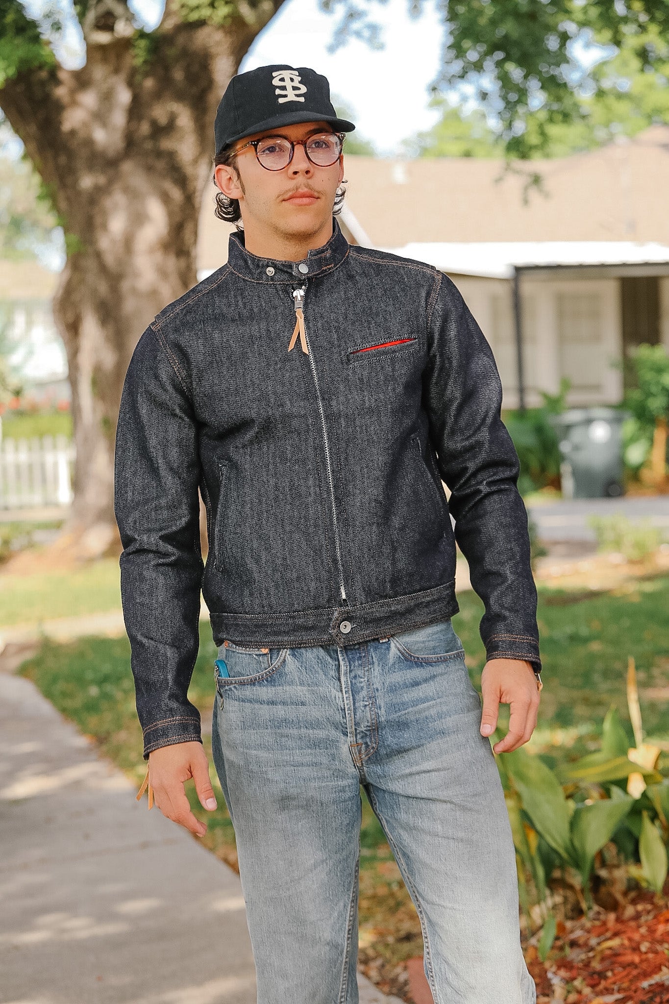 Person wearing a dark denim jacket and jeans standing outdoors with a house and trees in the background.