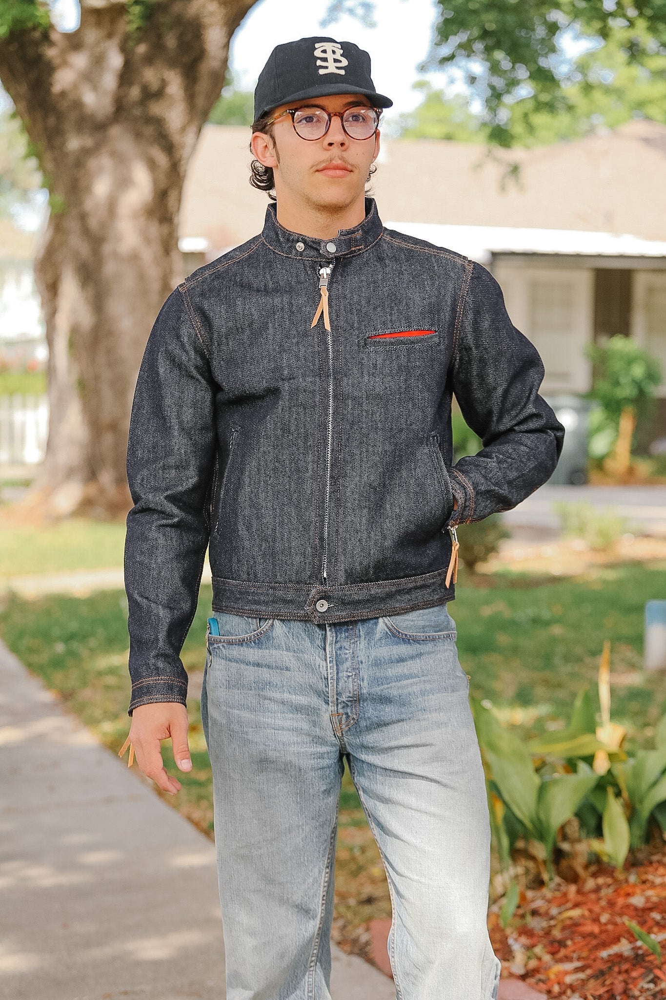 Man wearing a dark denim jacket and light jeans standing outdoors.