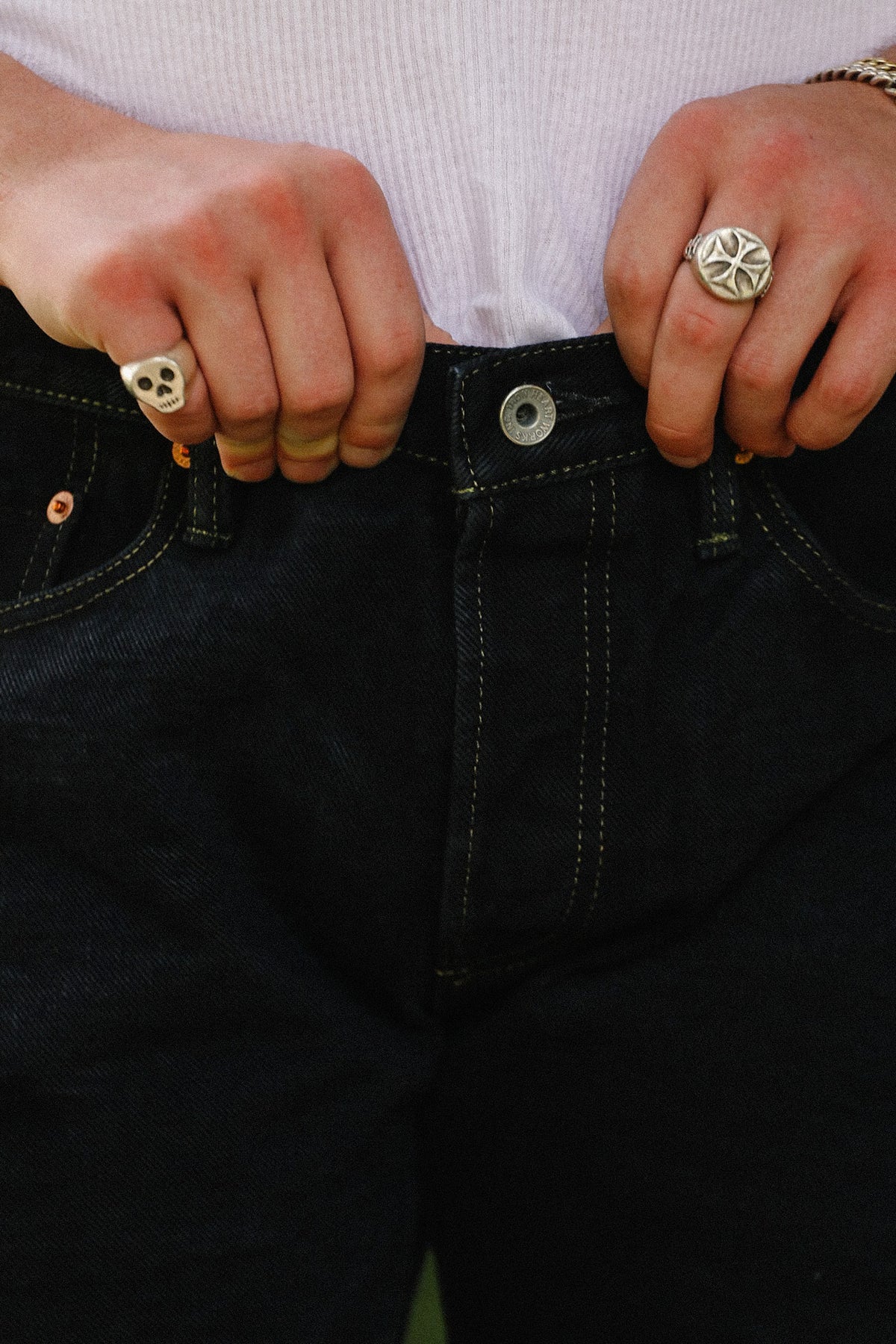 Close-up of a person wearing dark indigo jeans, pulling the waistband slightly outward with both hands. The person is also wearing silver skull and cross-shaped rings on their fingers.