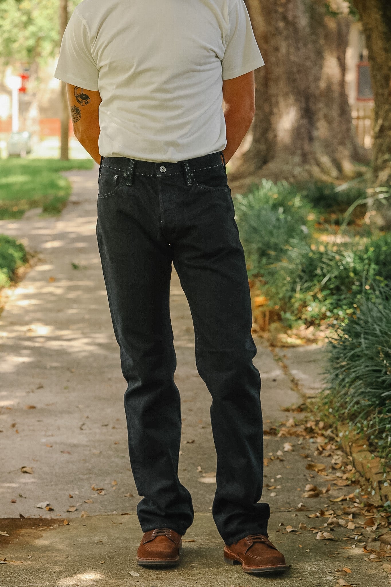 A person standing outdoors on a sidewalk wearing dark charcoal gray jeans, a white t-shirt, and brown shoes, photographed from the back.