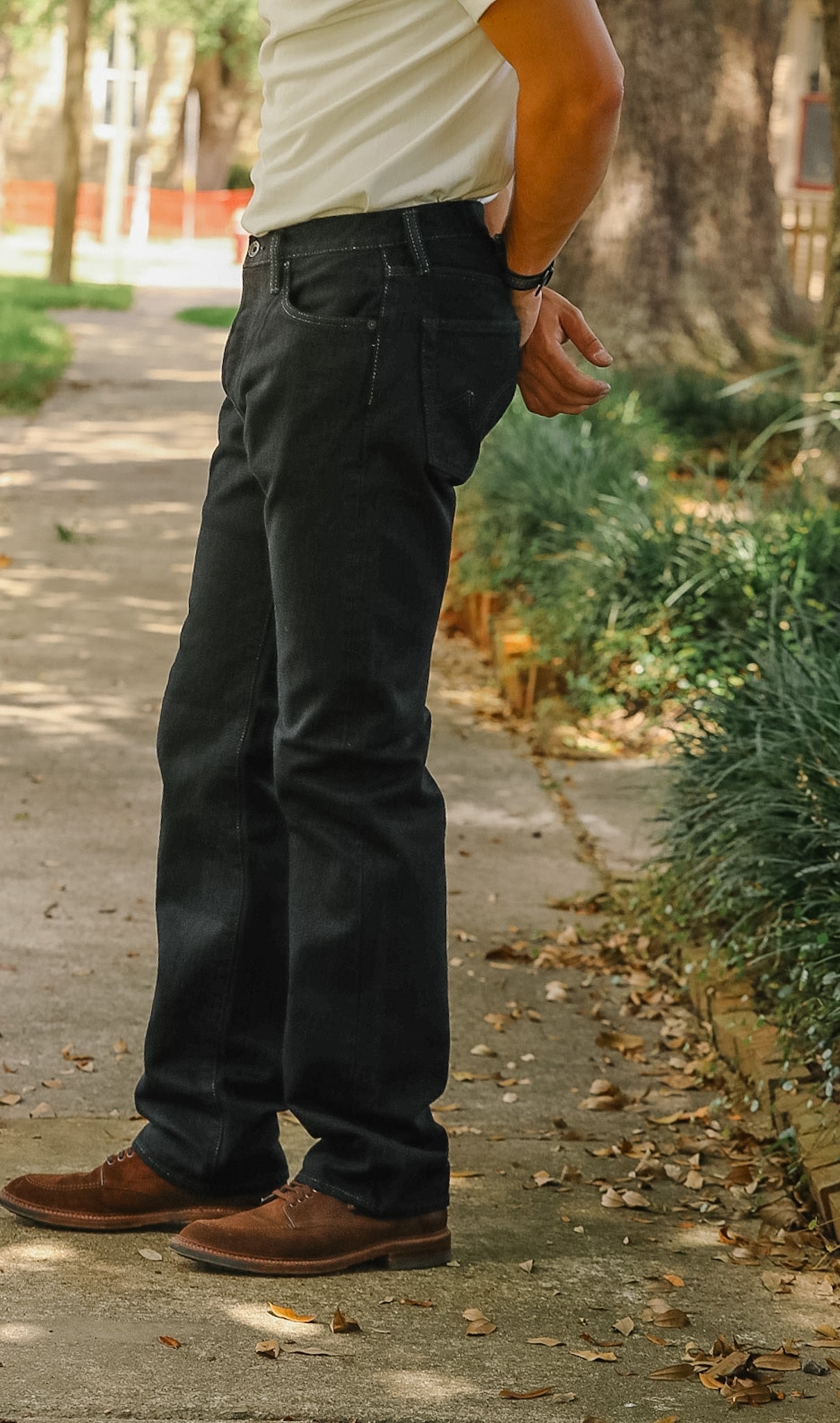 A person standing outdoors on a paved path wearing dark charcoal gray jeans, a light beige t-shirt, and brown leather shoes.