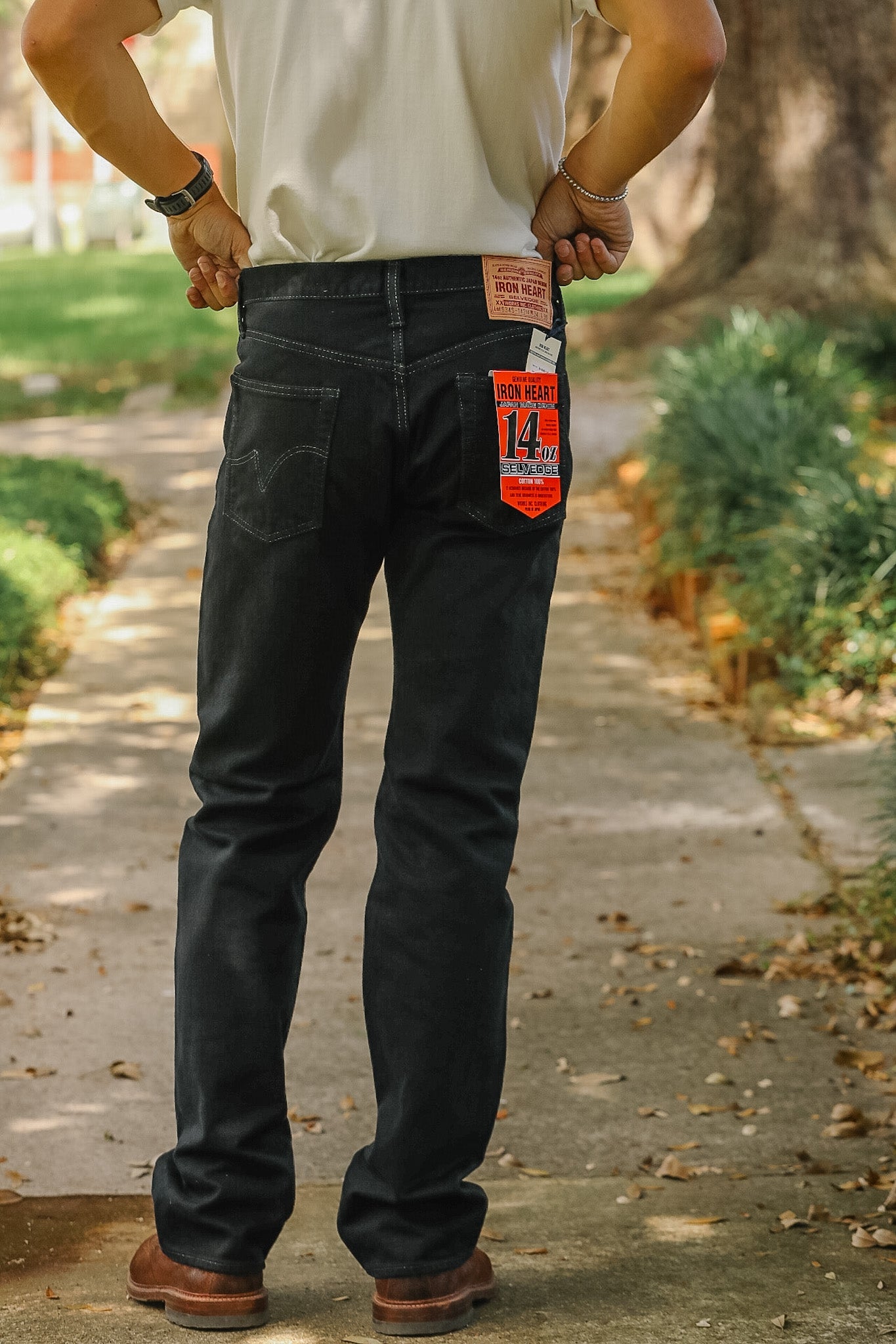 A man viewed from behind wearing dark wash denim jeans with a red and orange Levi's 501 label attached to the back pocket, standing on an outdoor paved path.