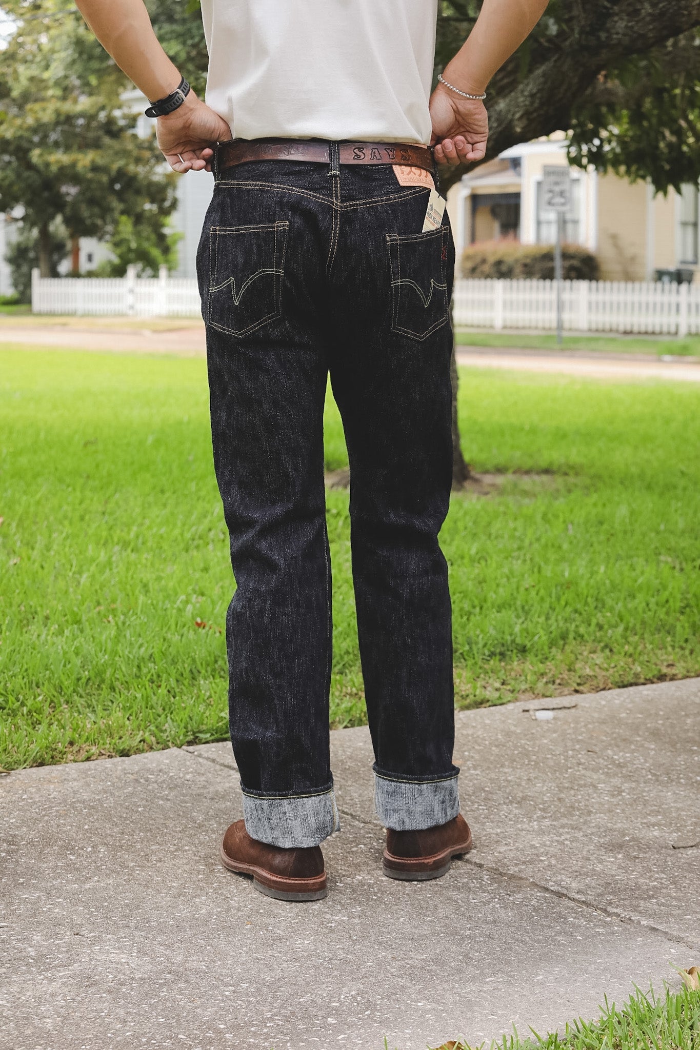 A person standing outdoors on a sidewalk wearing dark indigo denim jeans with rolled cuffs, a white shirt, and brown shoes, photographed from the back.