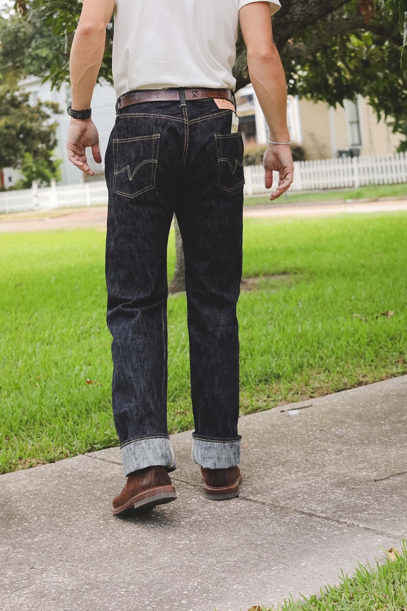 A person viewed from behind wearing dark indigo denim jeans with a rolled cuff, a white t-shirt, and brown boots while walking on a concrete sidewalk outdoors.