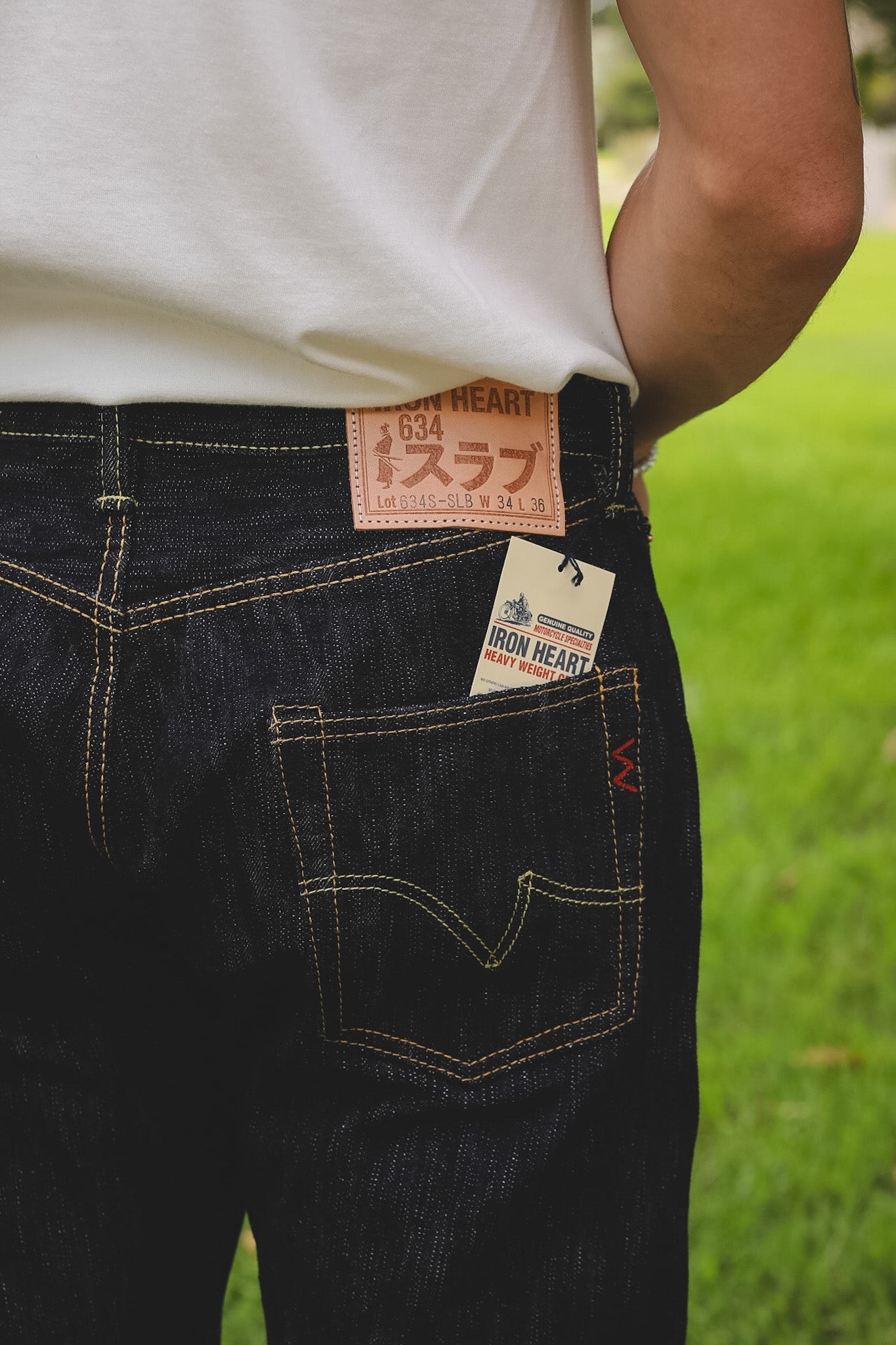 Close-up back view of a person wearing dark indigo denim jeans with a white t-shirt, showing the Iron Heart 634 label and hangtag on the waistband and back pocket.