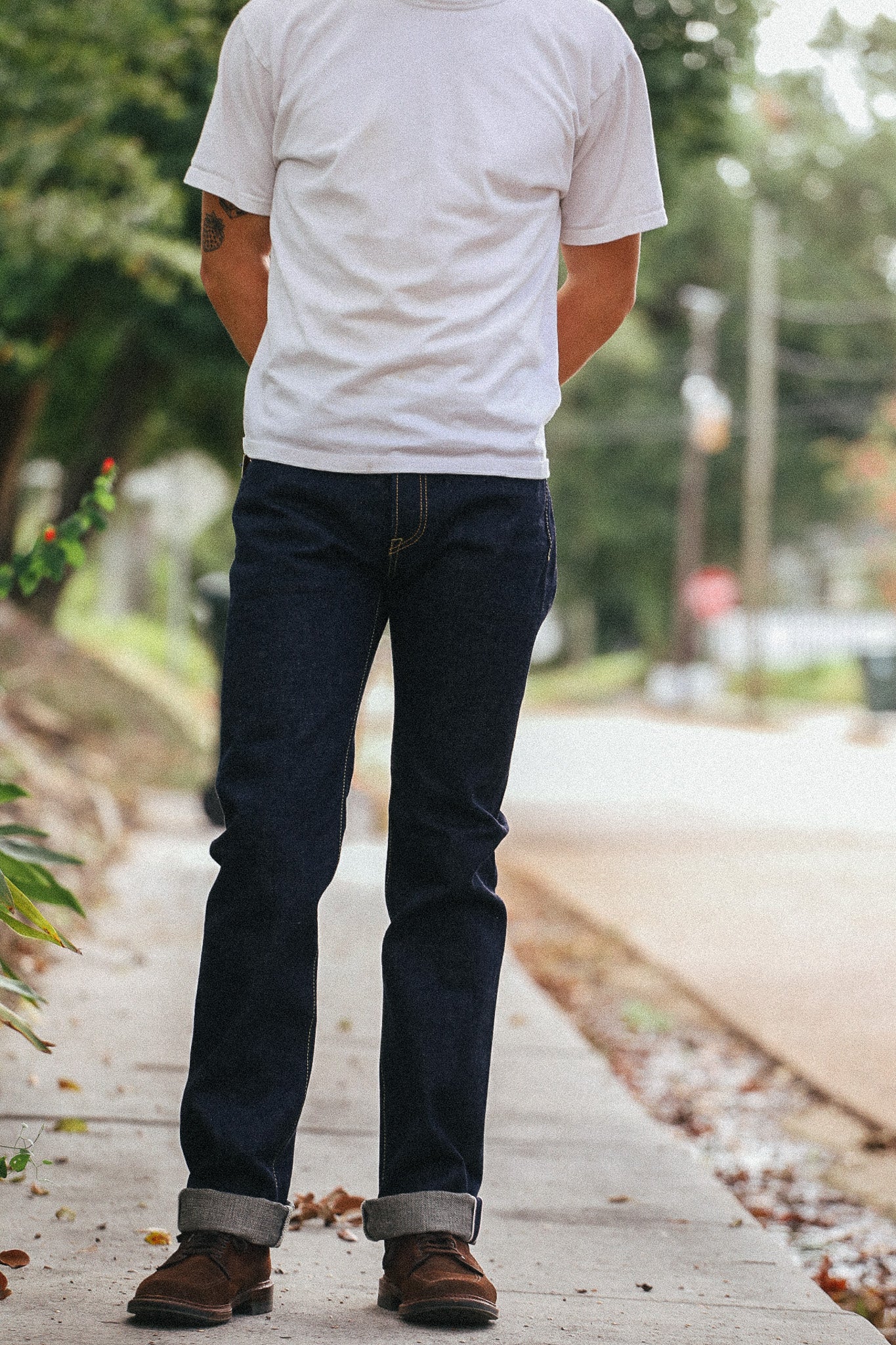 A person standing on a sidewalk outdoors, viewed from the back, wearing a plain white short-sleeve T-shirt, dark blue jeans with cuffed hems, and brown shoes.