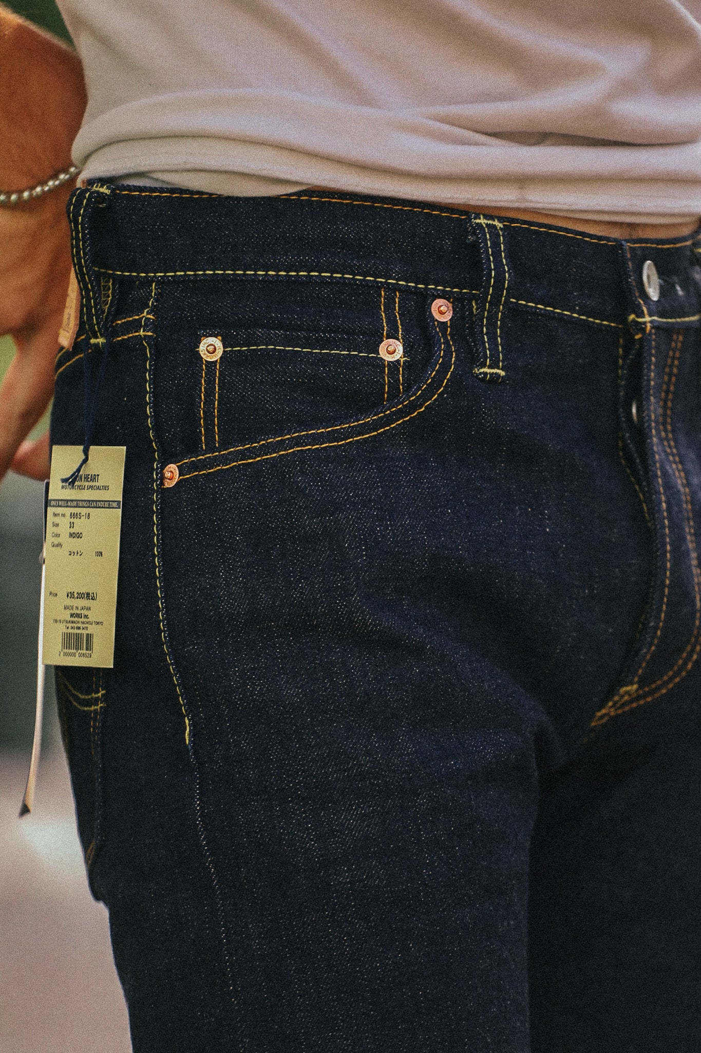 Close-up of a person wearing dark indigo denim jeans with yellow contrast stitching, a light beige t-shirt, and a paper product tag hanging from the pocket.