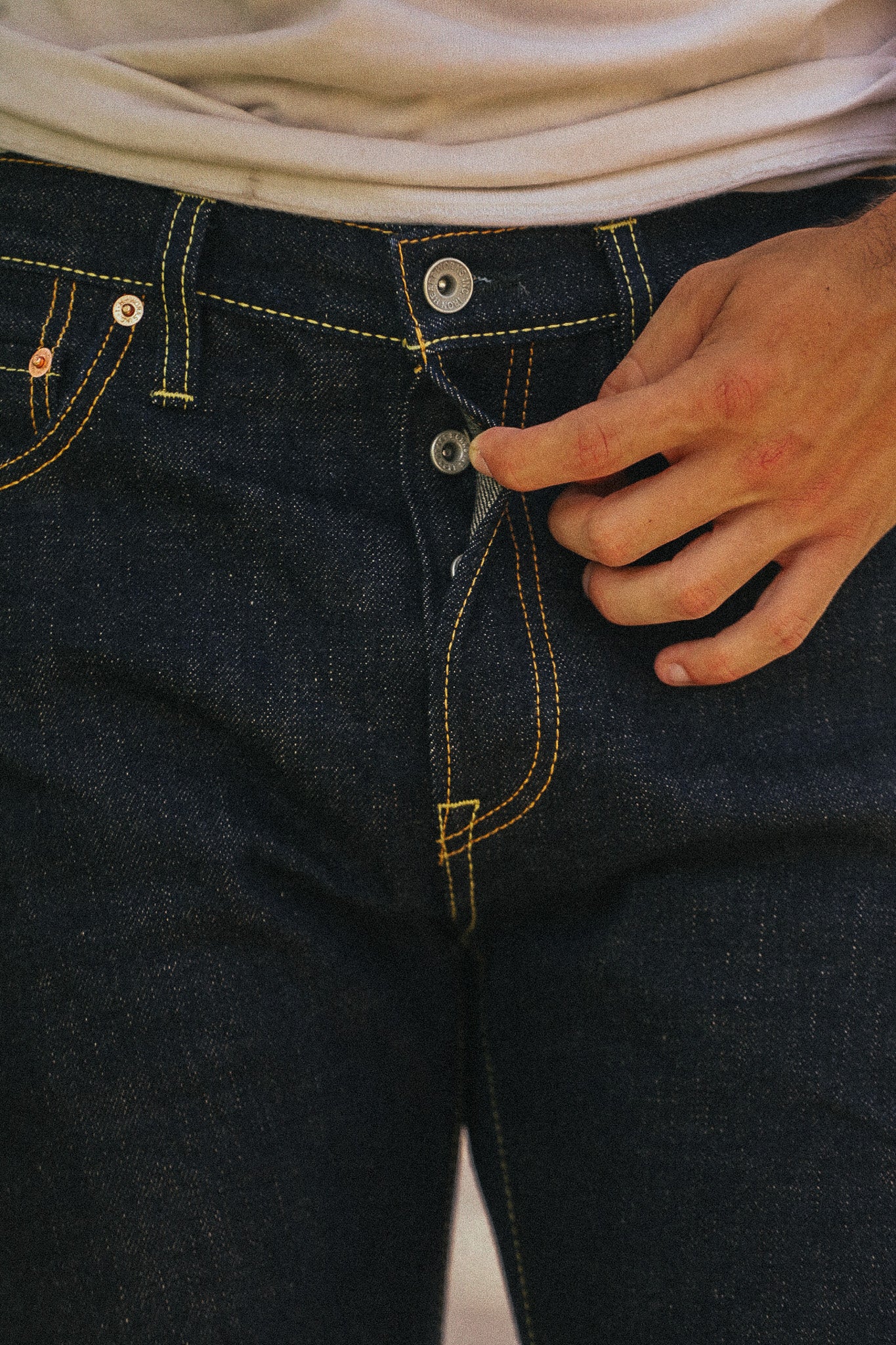 Close-up of a person wearing dark indigo denim jeans with yellow contrast stitching, pulling the front button closed with one hand.