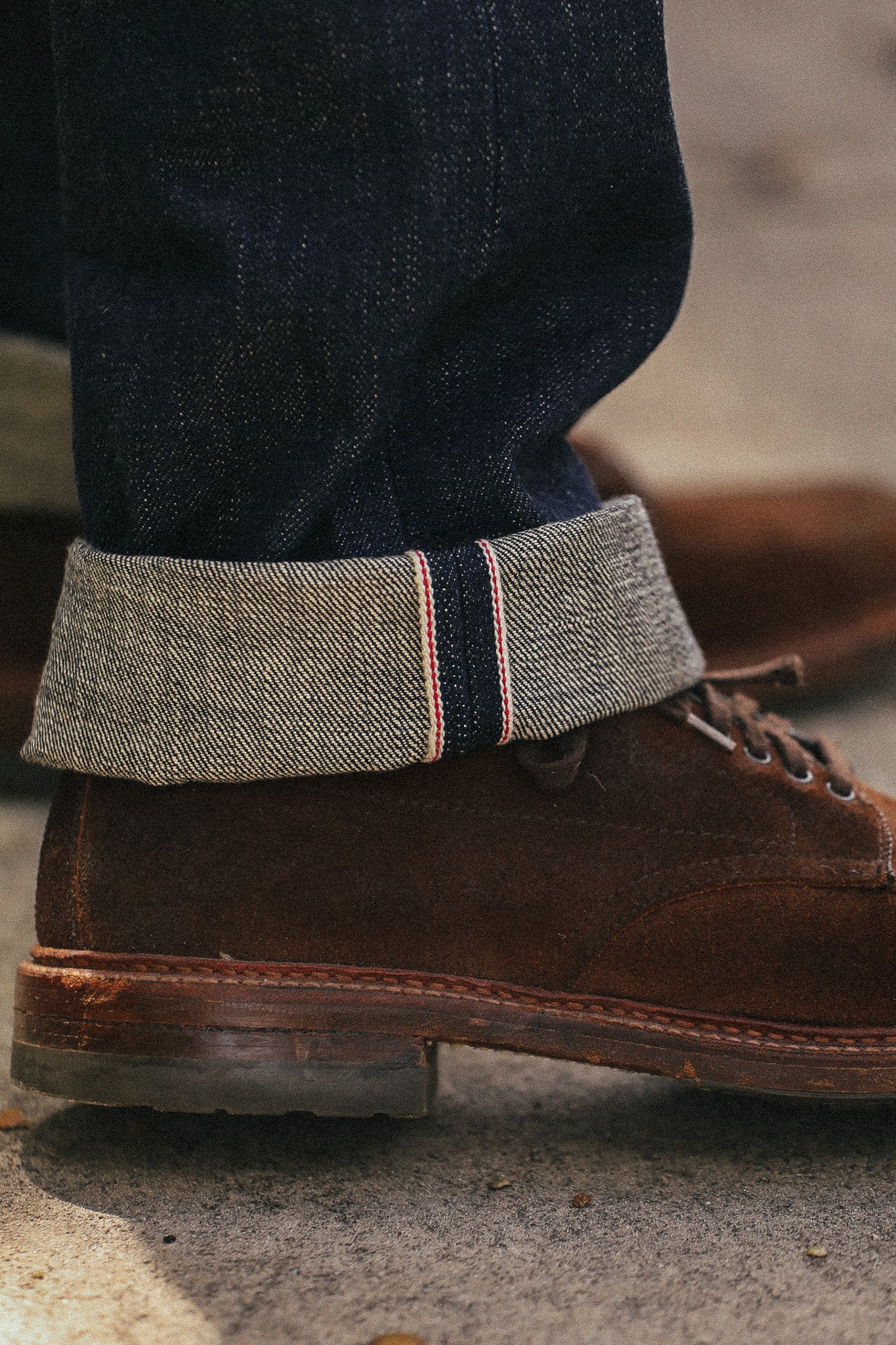 A close-up of a person's foot wearing a brown suede lace-up boot with dark blue jeans rolled up to show a textured gray herringbone fabric cuff with red and navy stripes.