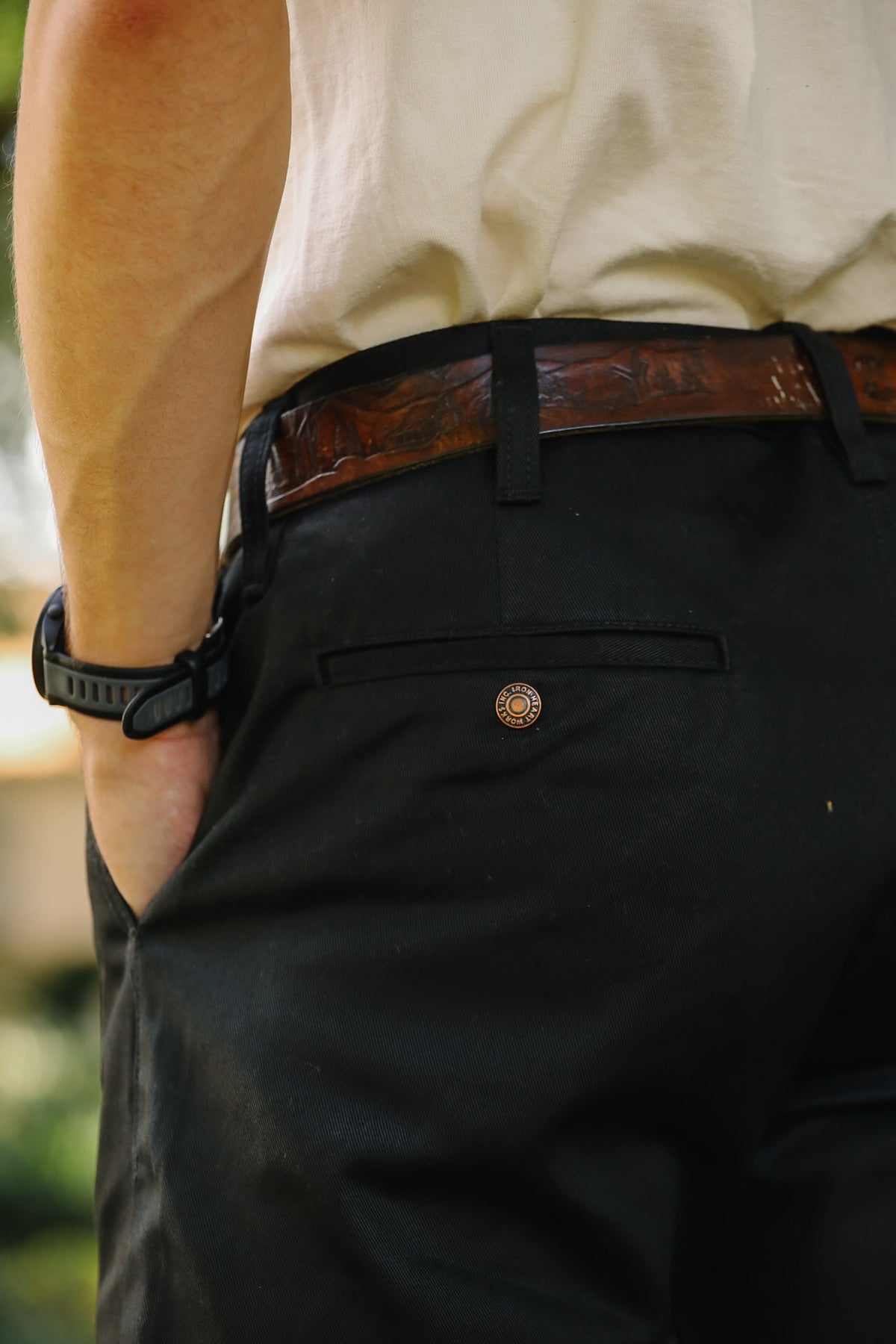 Close-up of a person wearing black dress pants with a brown leather embossed belt, a cream shirt, and a black wristwatch.