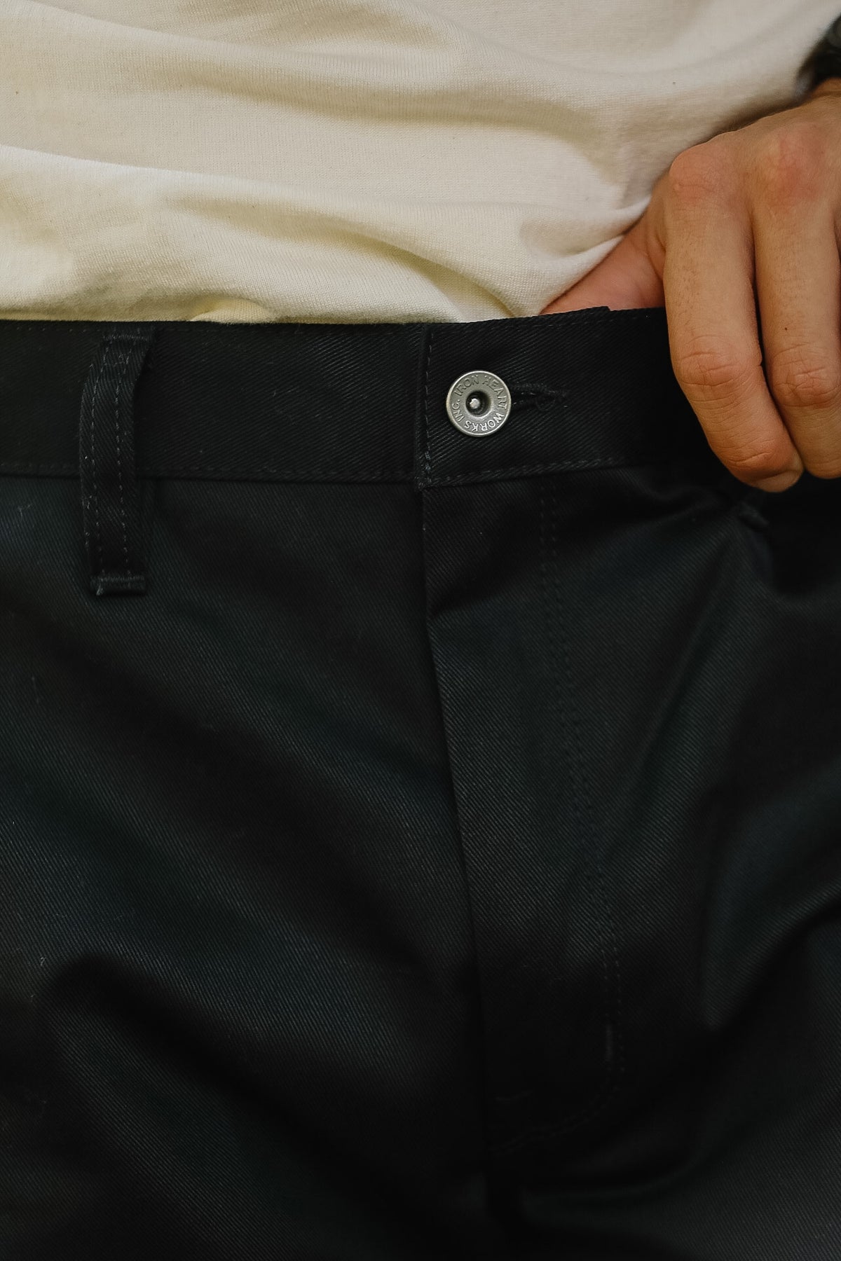 Close-up of a person wearing black trousers and a cream-colored shirt, with a hand pulling the waistband slightly to the side to demonstrate the button closure.