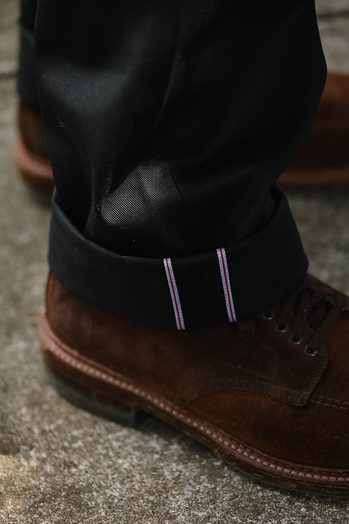 Close-up of a person's ankle wearing rolled-up black trousers with thin pink and white striped contrast stitching, paired with brown suede lace-up boots on a gray concrete surface.
