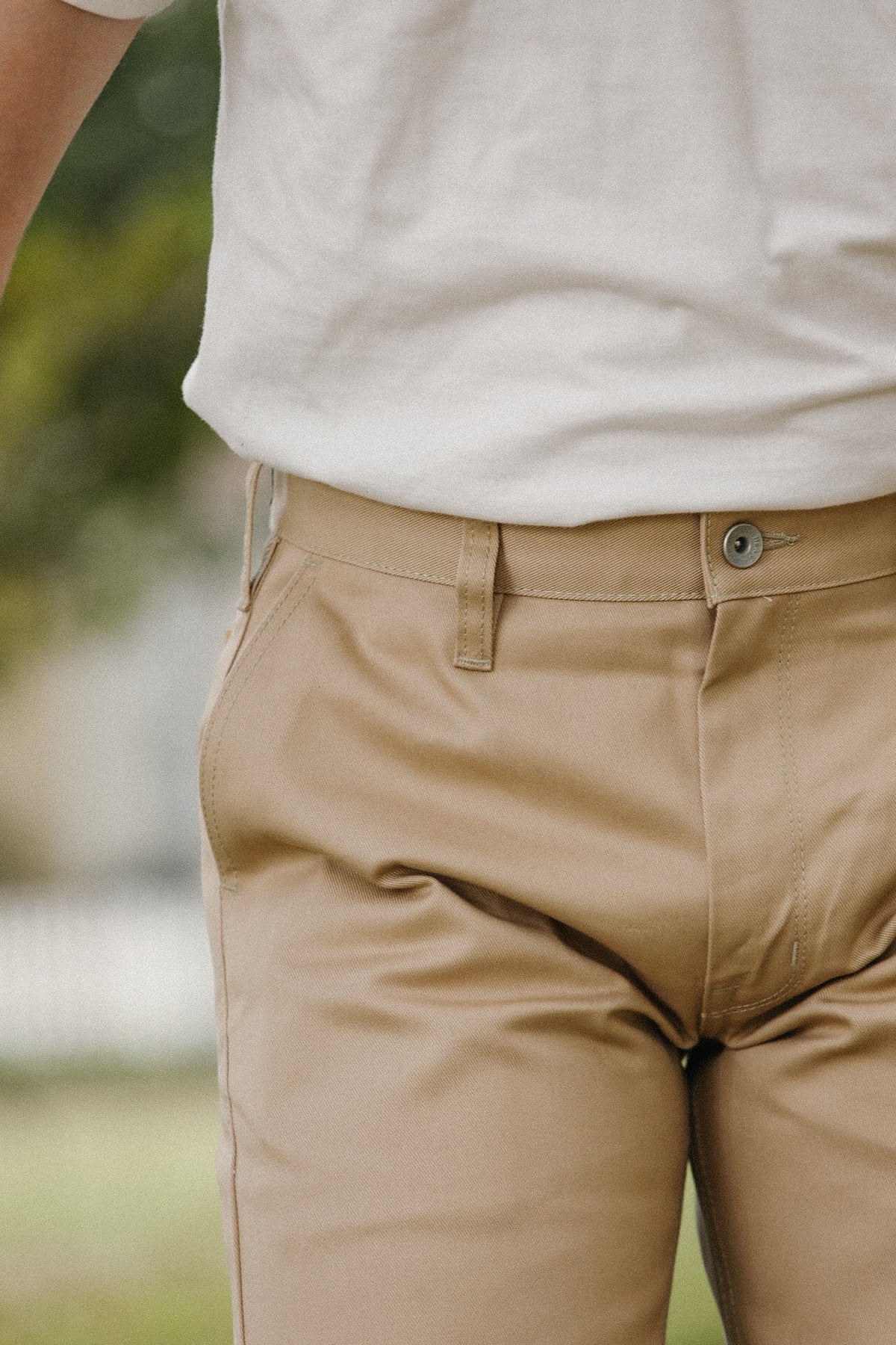Close-up of a person wearing tan chino pants and a white T-shirt, cropped from the chest down, with a softly blurred outdoor background.