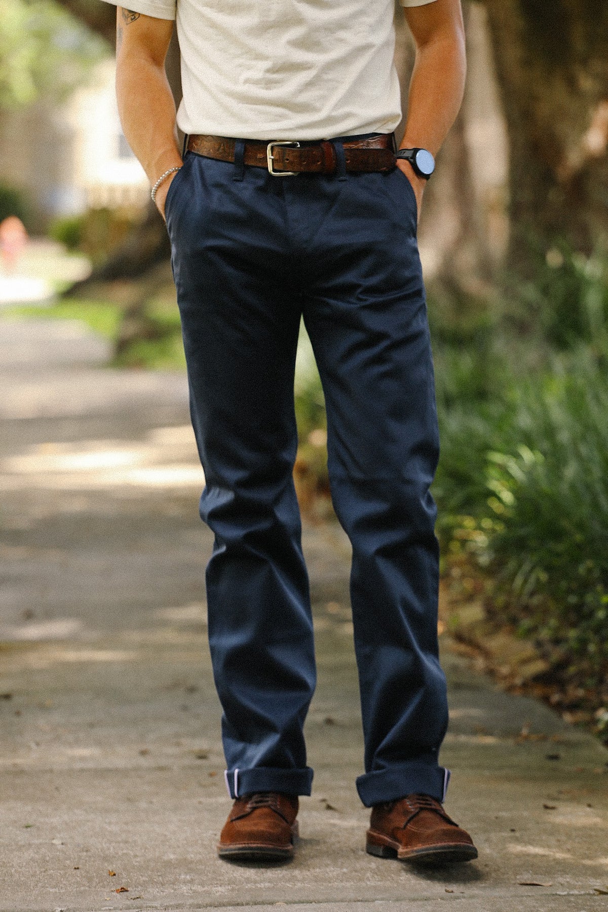 A person standing outdoors on a paved path wearing navy blue trousers, a brown belt, a white t-shirt, and brown shoes.