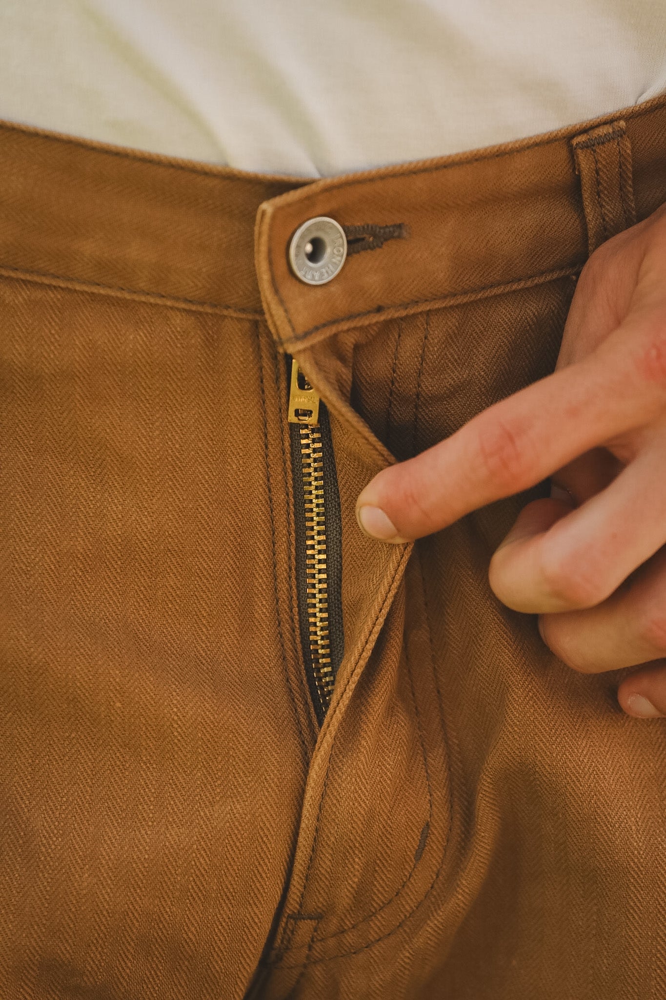 Close-up of a hand zipping up brown pants with a beige shirt underneath.