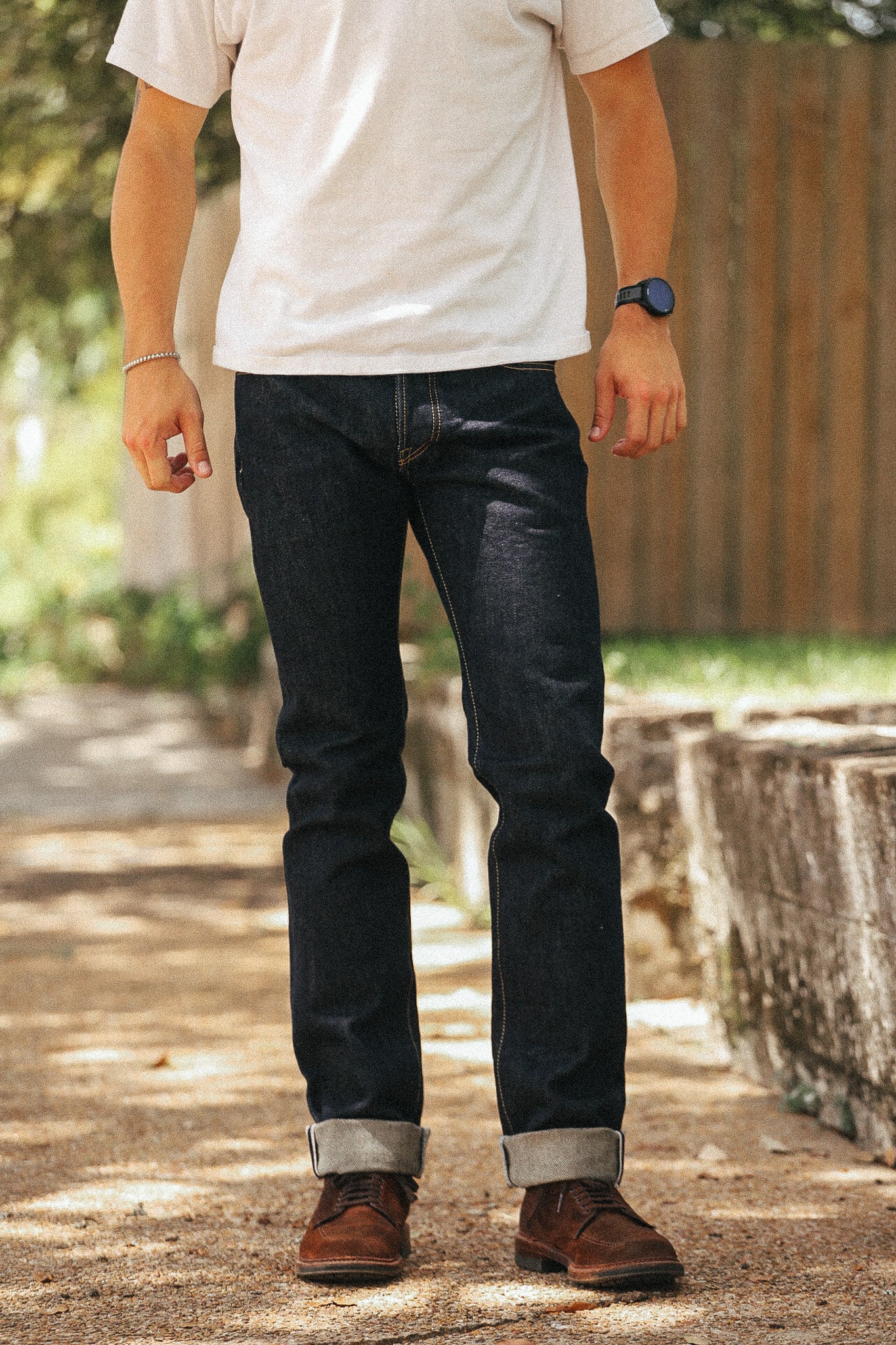 A person standing outdoors wearing dark indigo denim jeans with rolled cuffs, a white t-shirt, and brown leather shoes.